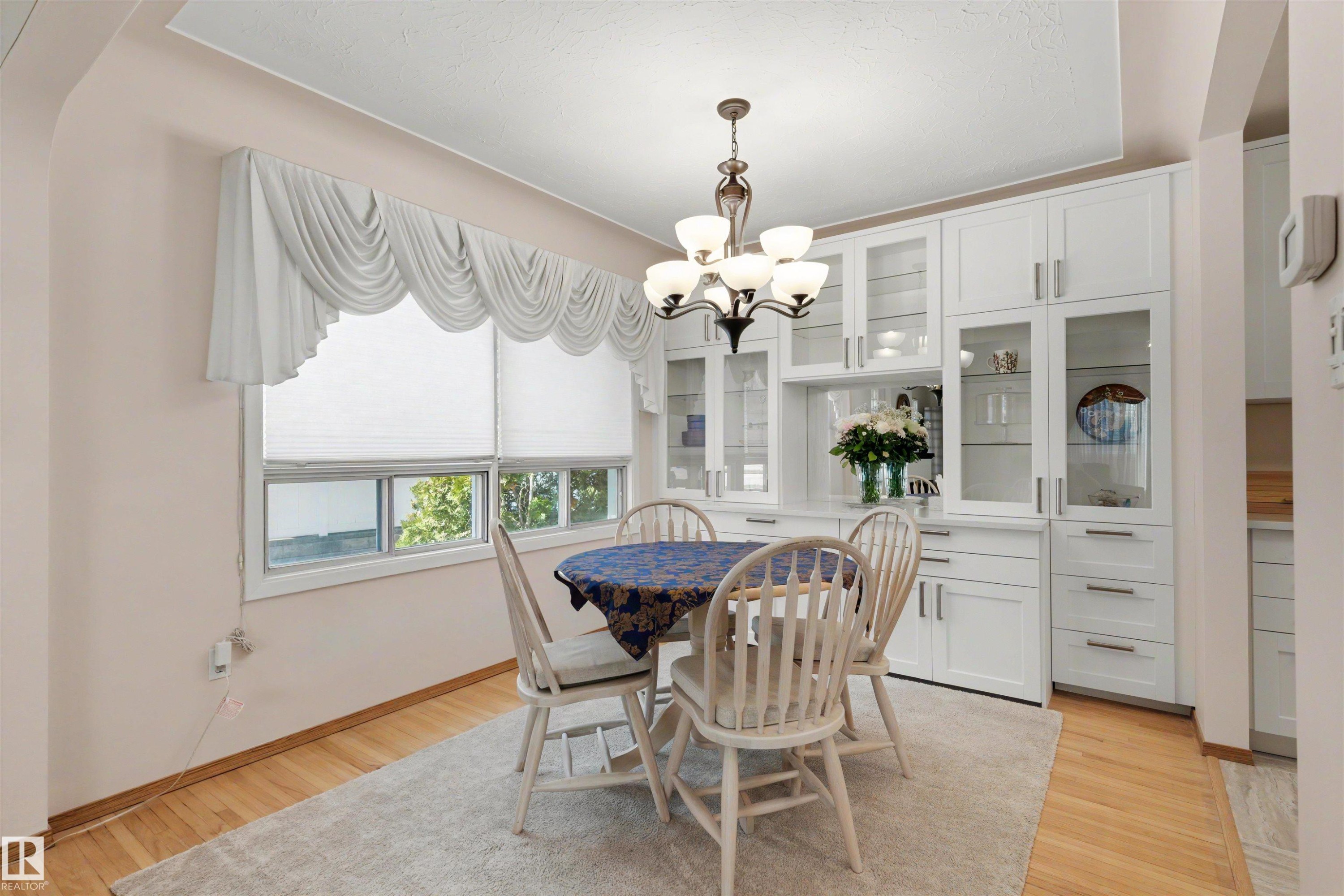 Dining area featuring extensive built-in cabinetry with glass display fronts, wood-finish flooring, a contemporary chandelier, and a window with white blinds - 9602 85 Street, Edmonton, AB