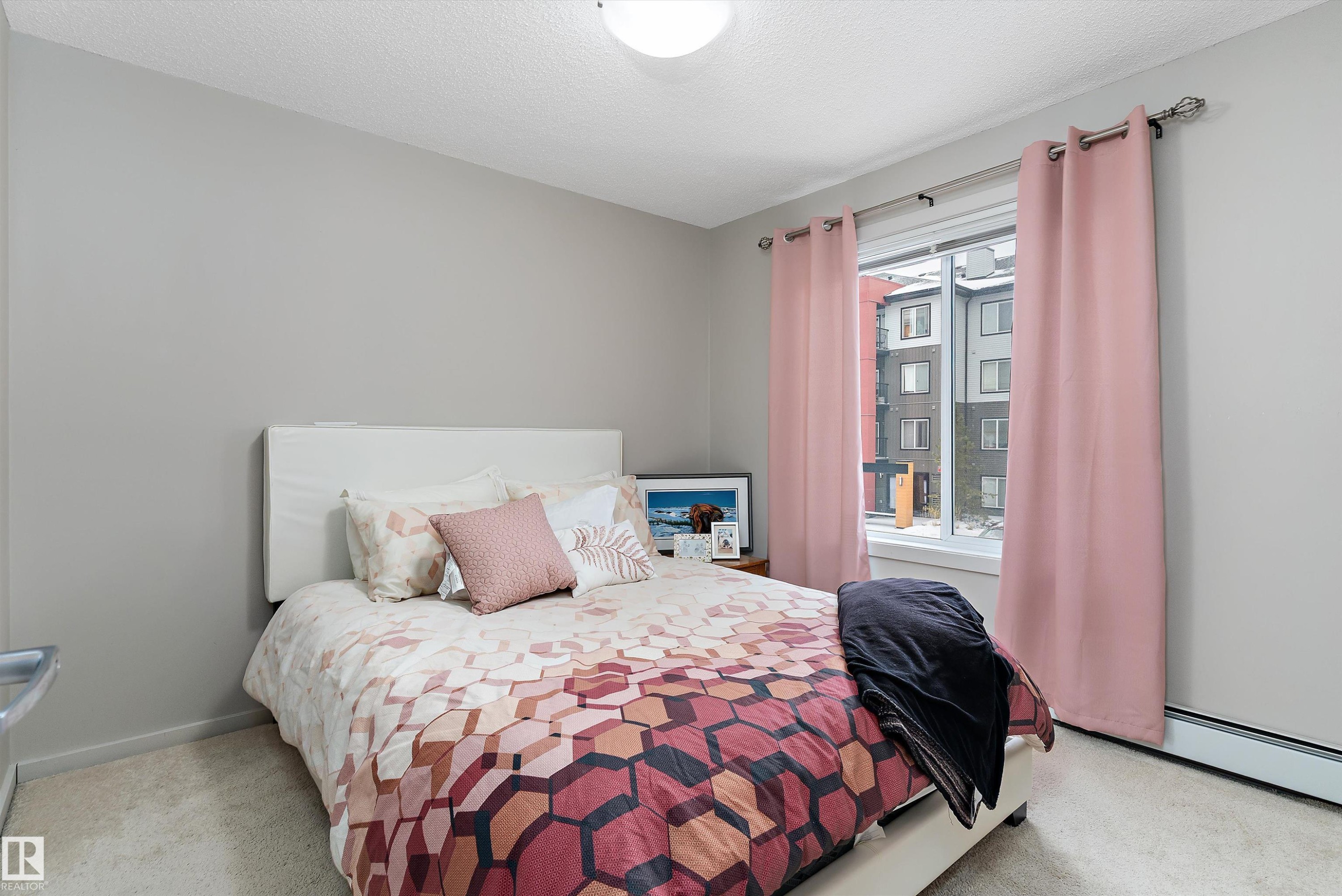 Carpeted bedroom with a window featuring a white frame and pink curtains, light gray walls, and a ceiling-mounted light fixture - 231 344 Windermere Road, Edmonton, AB - Indoor Photo Showing Bedroom