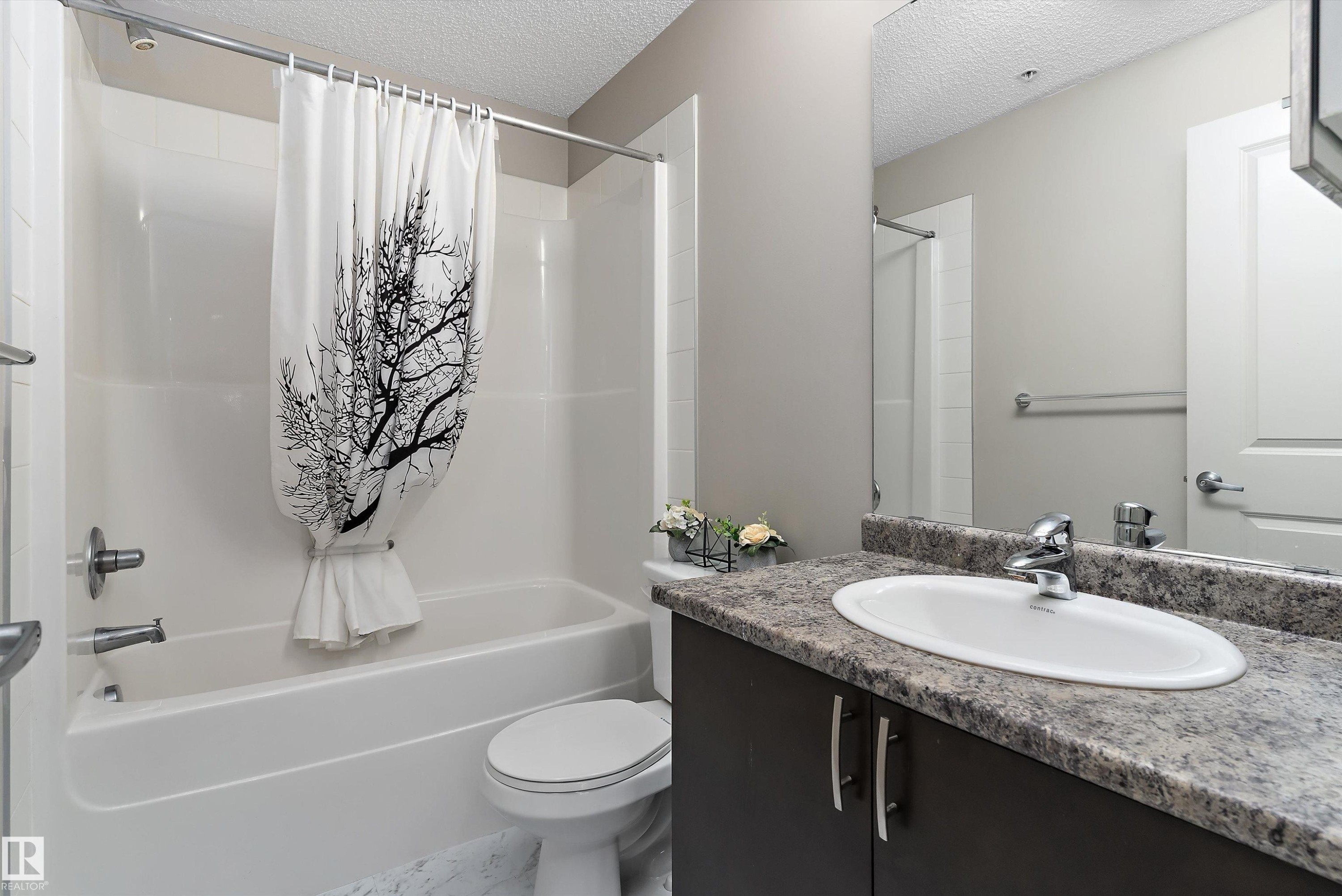 Bathroom featuring a white bathtub and shower combination, a dark wood-finish vanity with a speckled countertop, an oval undermount sink, and a large wall-mounted mirror - 231 344 Windermere Road, Edmonton, AB - Indoor Photo Showing Bathroom