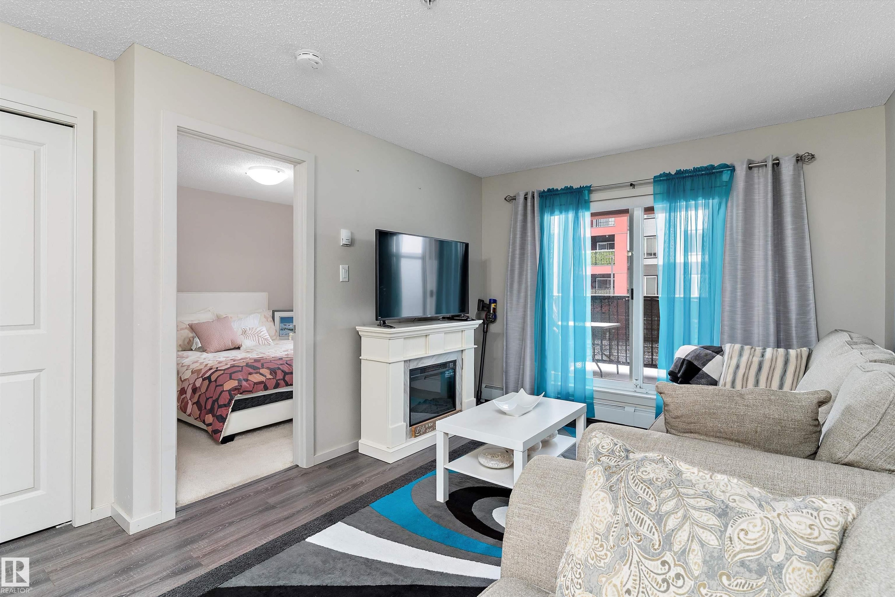 Living area featuring wood-finish flooring, a built-in electric fireplace with mantel, and a balcony with sliding glass doors - 231 344 Windermere Road, Edmonton, AB - Indoor Photo Showing Other Room With Fireplace