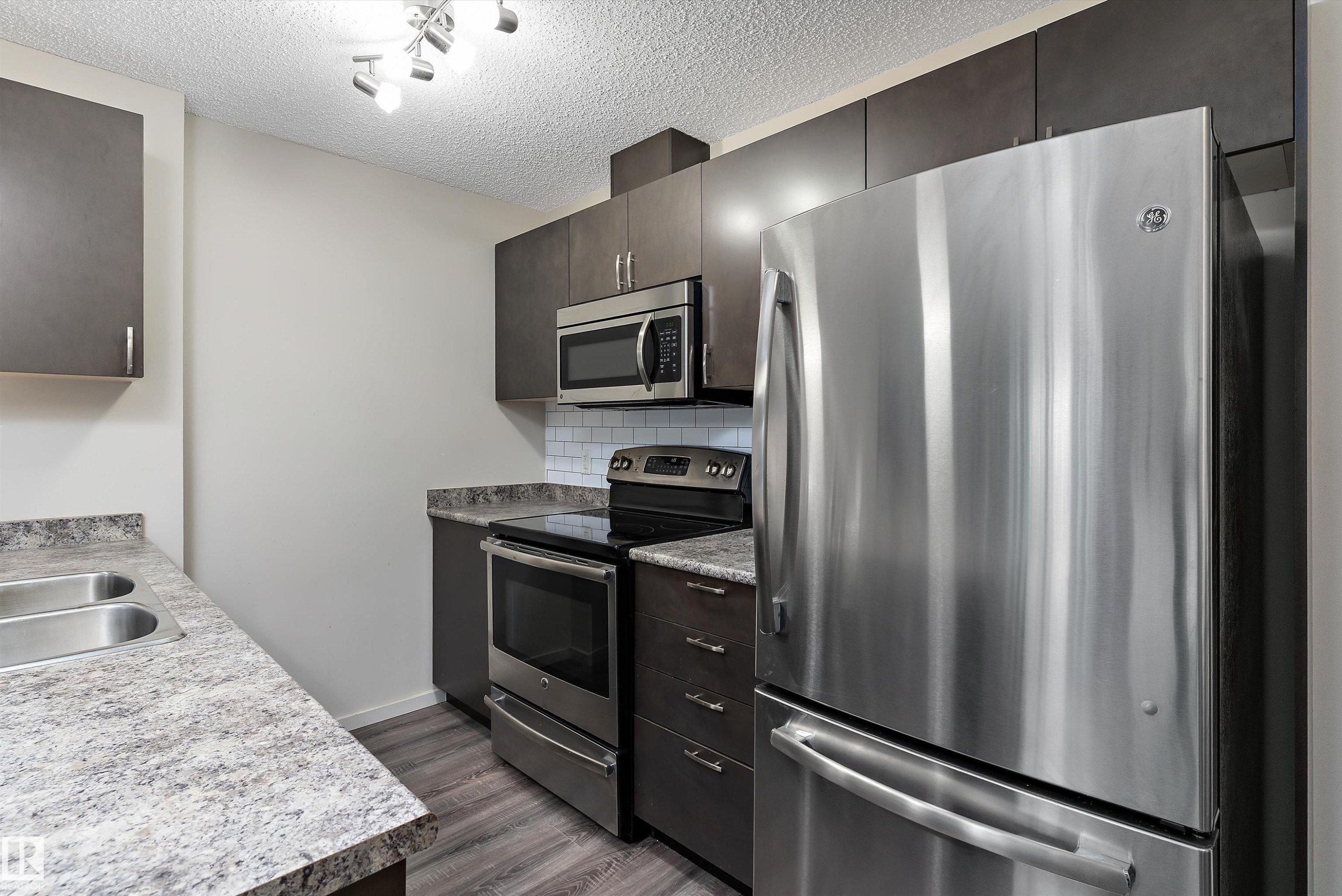 Contemporary kitchen featuring dark wood-finish cabinetry, stainless steel appliances, a subway tile backsplash, and wood-finish flooring - 231 344 Windermere Road, Edmonton, AB - Indoor Photo Showing Kitchen With Double Sink