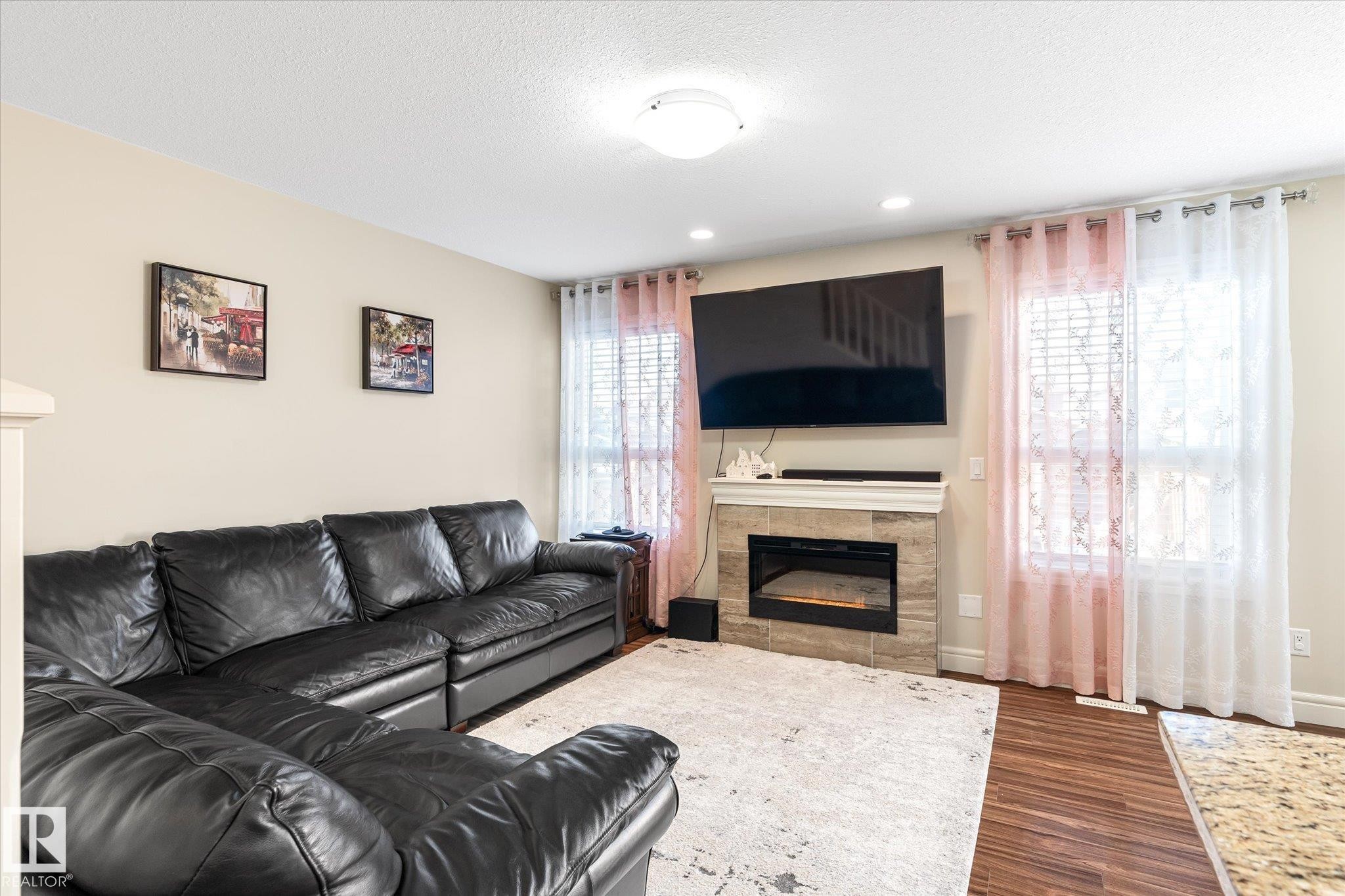 Living area featuring an electric fireplace with a tile surround and mantel, wood-finish flooring, two windows with blinds, and recessed ceiling lighting - 6864 Evans Wynd, Edmonton, AB - Indoor Photo Showing Living Room With Fireplace