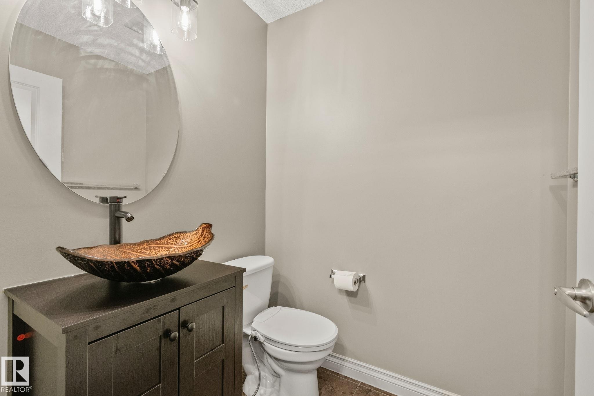 Powder room featuring a dark wood-finish vanity with a vessel sink and a wall-mounted faucet, complemented by an oval mirror and a three-light vanity fixture - 46 445 Brintnell Boulevard, Edmonton, AB - Indoor Photo Showing Bathroom