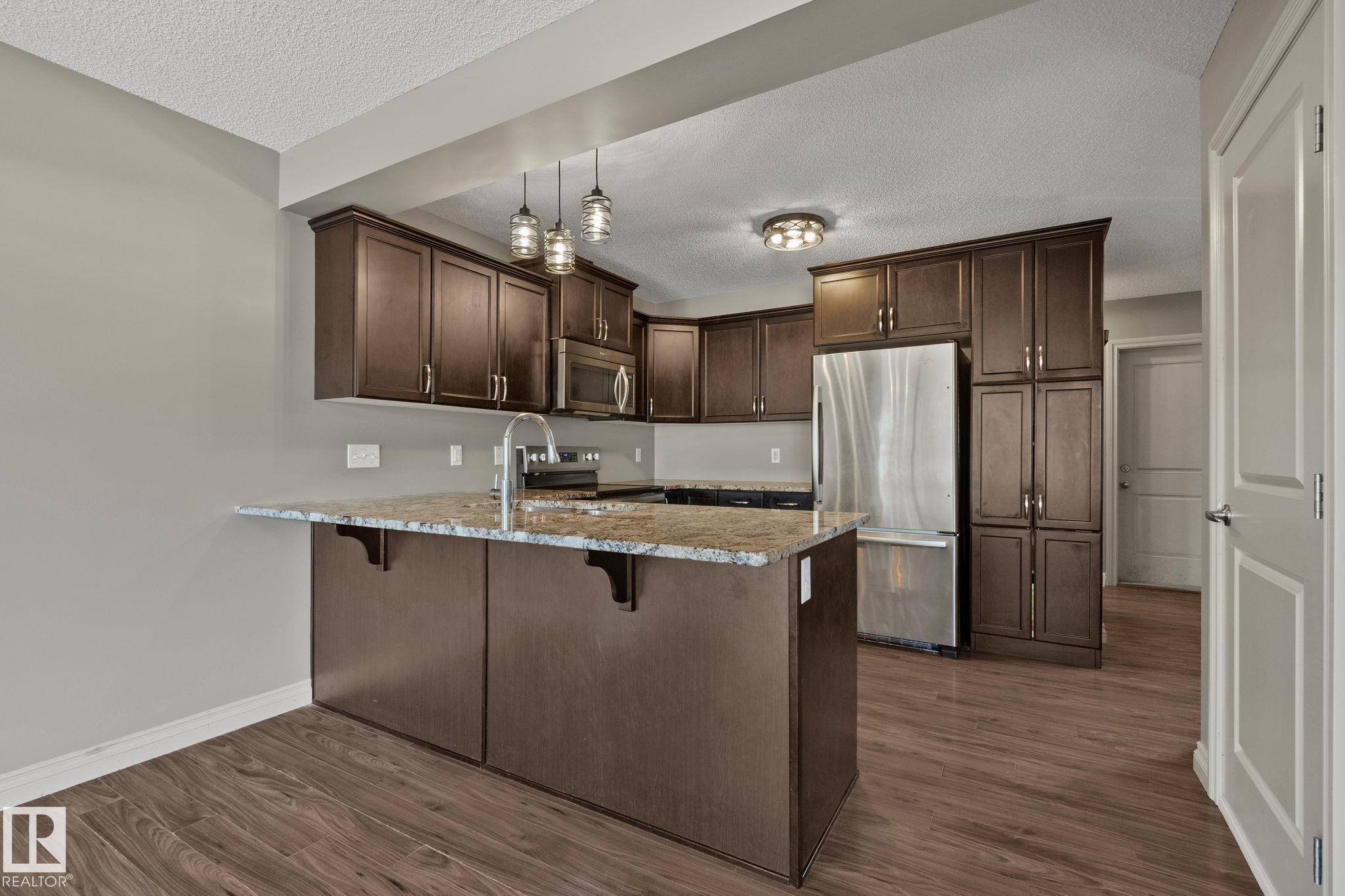 Kitchen featuring an island with stone-finish countertop, dark wood-finish cabinetry, stainless steel appliances, and wood-finish flooring - 46 445 Brintnell Boulevard, Edmonton, AB - Indoor Photo Showing Kitchen With Stainless Steel Kitchen