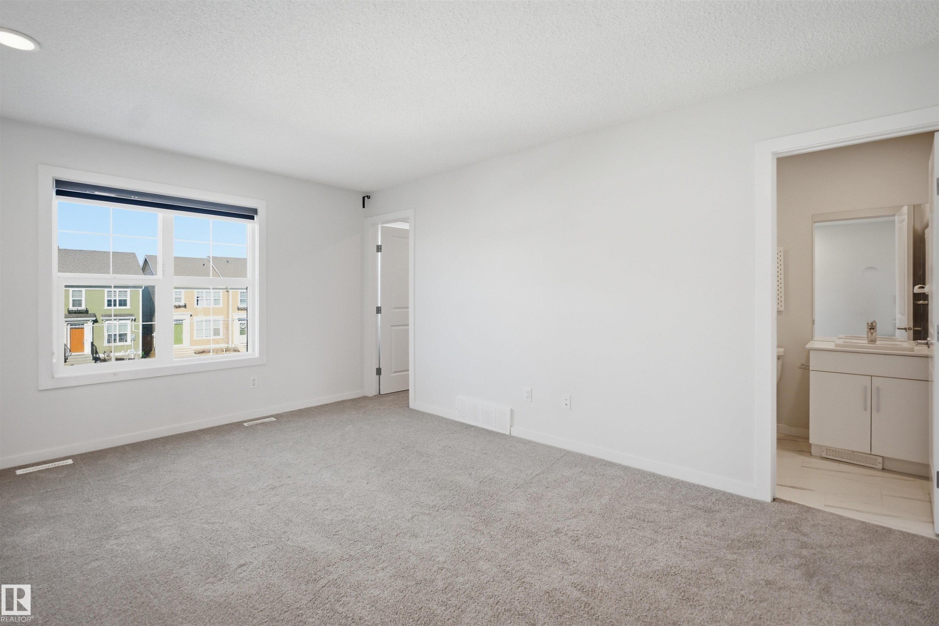 This spacious room features light gray carpeting, neutral white walls, a large window, and an en-suite bathroom with a vanity and mirror - 2125 Maple Road Nw, Edmonton, AB - Indoor Photo Showing Other Room