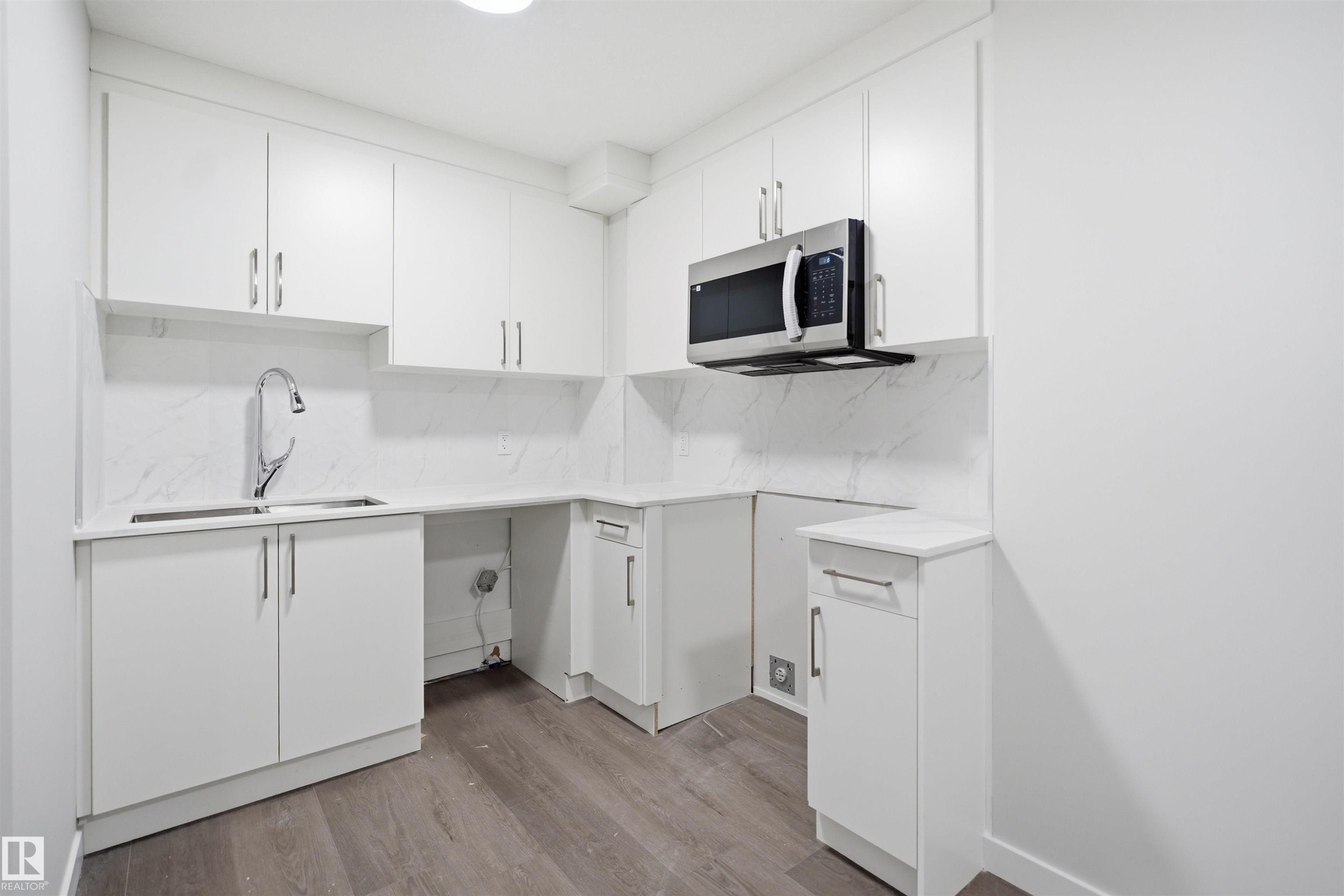 The kitchen features white cabinetry, a double basin stainless steel sink with a gooseneck faucet, a built-in microwave, and wood-style flooring - 2125 Maple Road Nw, Edmonton, AB - Indoor Photo Showing Kitchen