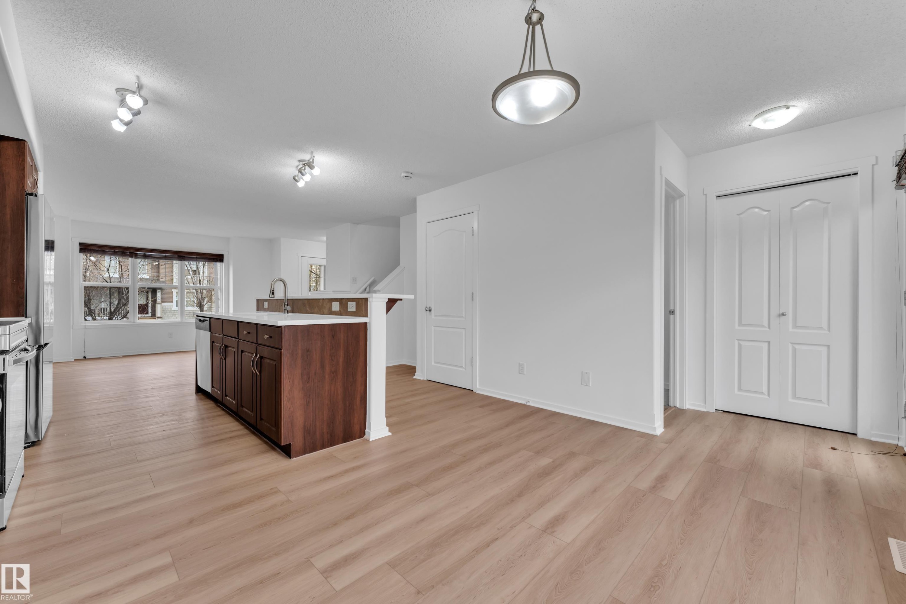 Open-concept kitchen and living area featuring wood-finish flooring, a kitchen island with dark wood cabinetry and light countertops, stainless steel appliances, and a large window with horizontal blinds - 2143 32 Street, Edmonton, AB