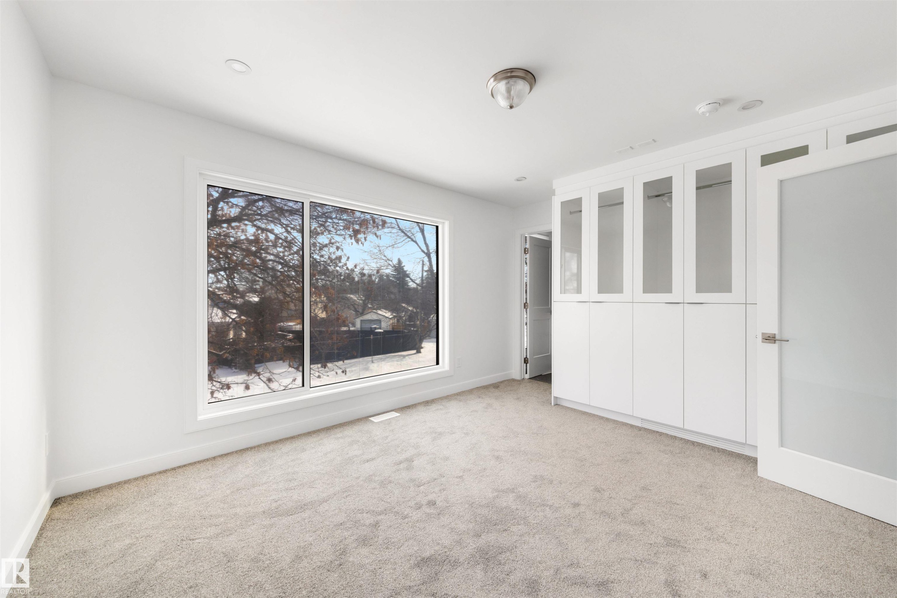 This room features a large window, light-colored carpeting, and a built-in white storage unit with glass-fronted upper cabinets - 7927 112S Avenue, Edmonton, AB - Indoor Photo Showing Other Room