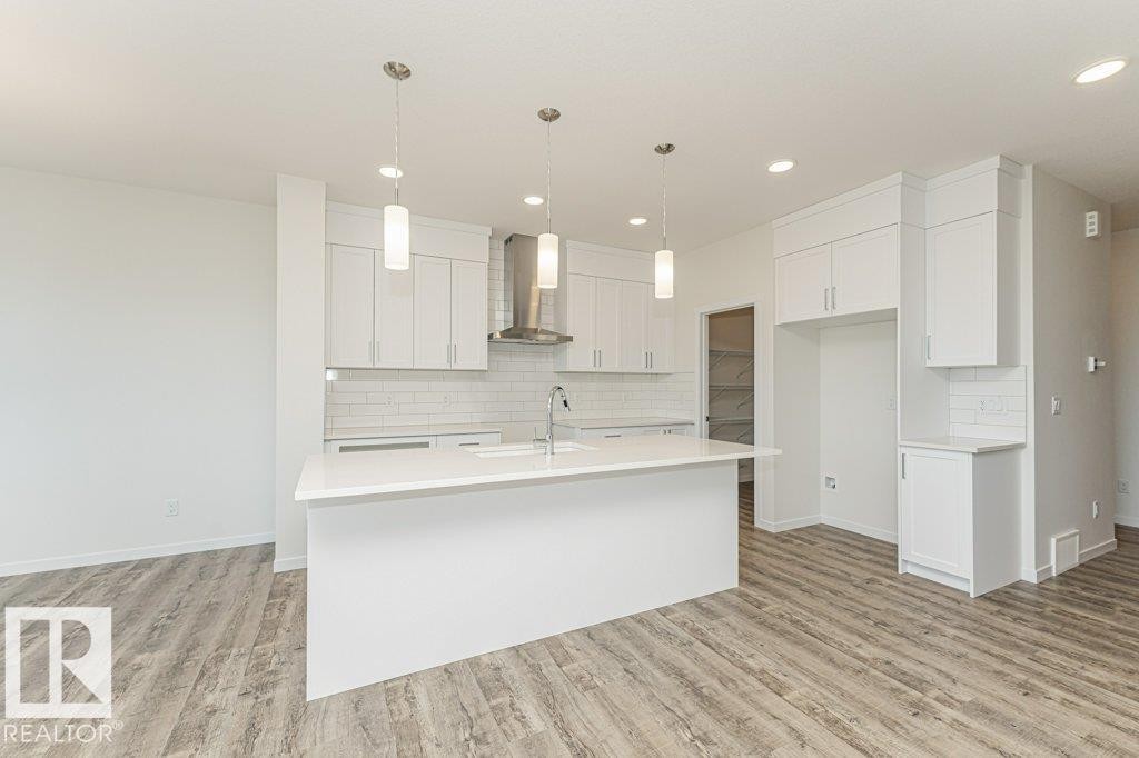 Modern kitchen featuring white cabinetry, a large center island with integrated sink, stainless steel range hood, white subway tile backsplash, and wood-finish flooring - 6271 180 Avenue Nw, Edmonton, AB