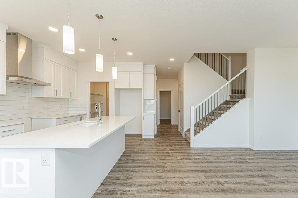 Modern kitchen featuring an expansive island with an undermount sink, white shaker cabinetry, stainless steel range hood, and subway tile backsplash - 6271 180 Avenue Nw, Edmonton, AB