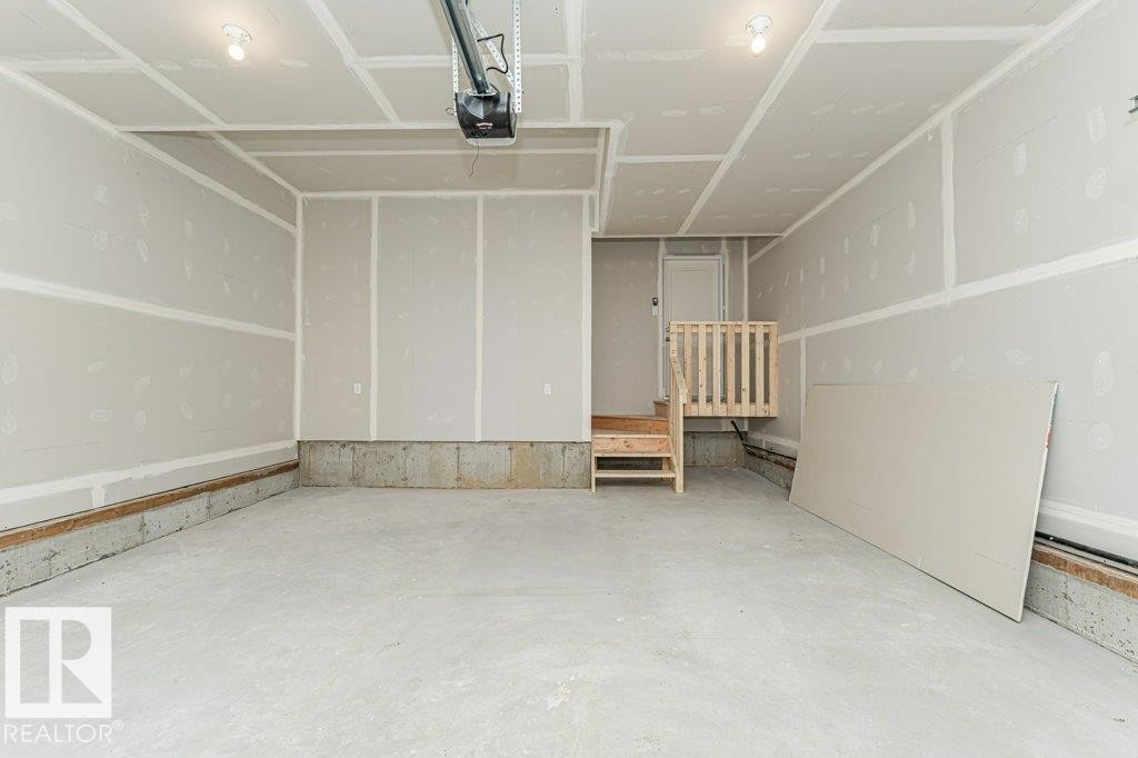 Unfinished garage interior featuring drywall-finished walls and ceiling, a concrete floor, and a wooden stairwell with railing - 6271 180 Avenue Nw, Edmonton, AB