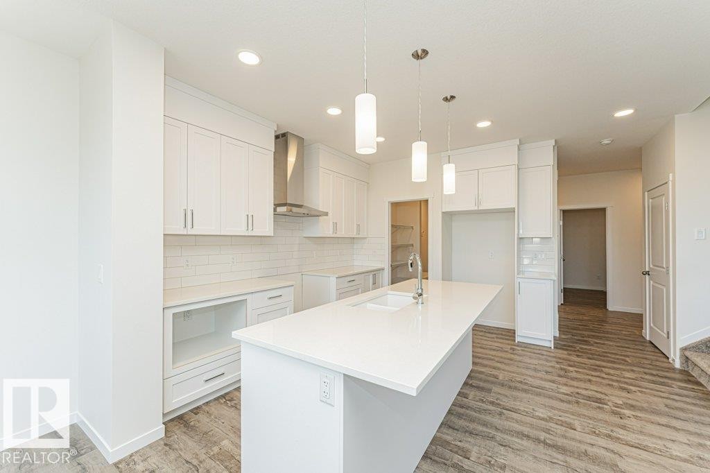 Modern kitchen featuring a large center island with integrated sink, white cabinetry, subway tile backsplash, and wood-finish flooring - 6271 180 Avenue Nw, Edmonton, AB