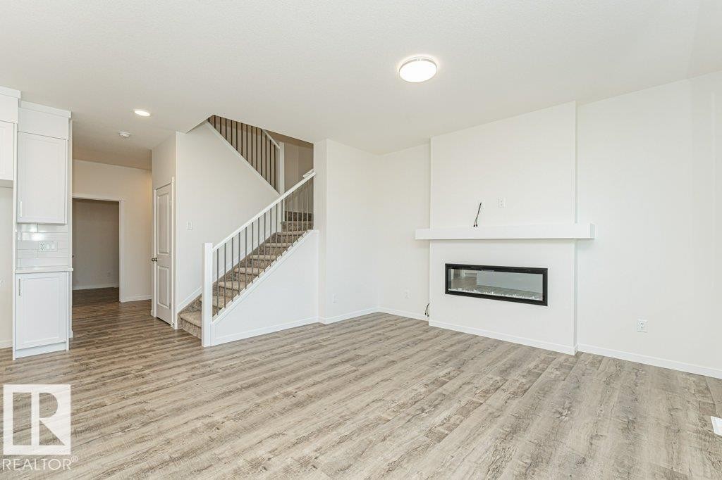Living area featuring wood-finish flooring, a contemporary fireplace with a white mantel, and a staircase with carpeted treads and white railings - 6271 180 Avenue Nw, Edmonton, AB