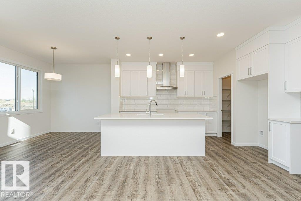 Bright kitchen featuring white cabinetry, a central island with a sink, stainless steel appliances including a range hood, and a white subway tile backsplash - 6271 180 Avenue Nw, Edmonton, AB