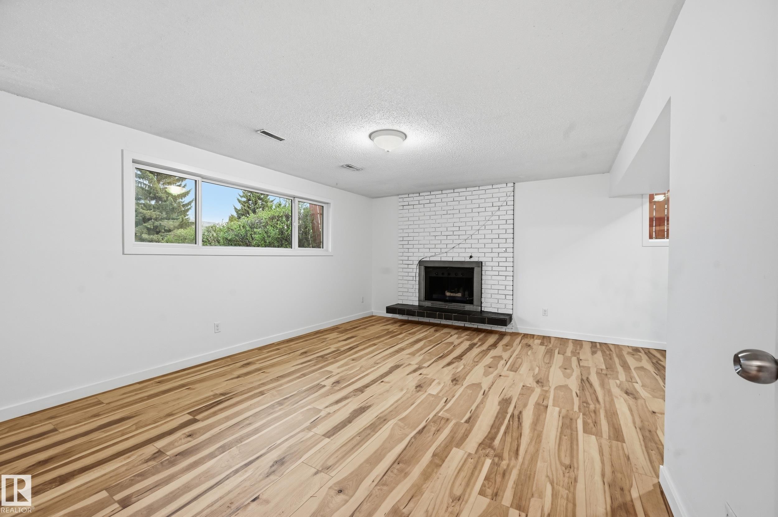 Spacious room featuring wood-finish flooring, a white brick fireplace with a dark hearth, and a large multi-pane window with exterior landscaping - 13903 87 Street, Edmonton, AB - Indoor Photo Showing Other Room With Fireplace