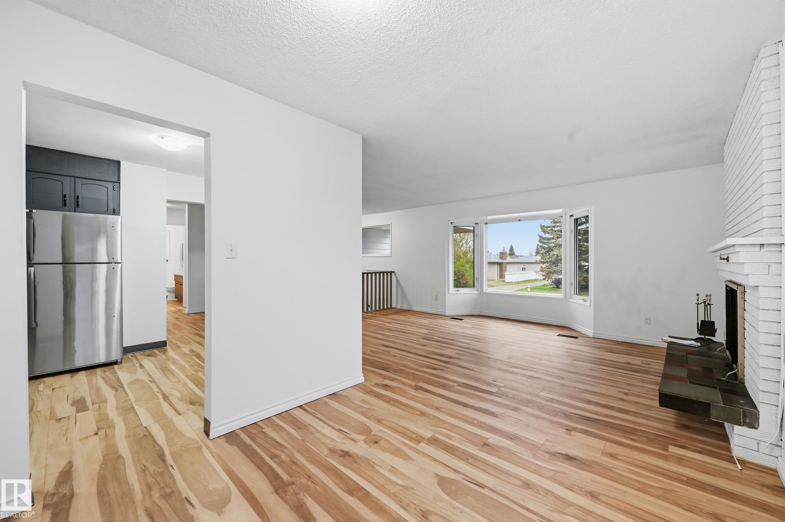 Bright living area featuring wood-finish flooring, a white brick fireplace with hearth, and a large bay window - 13903 87 Street, Edmonton, AB - Indoor With Fireplace