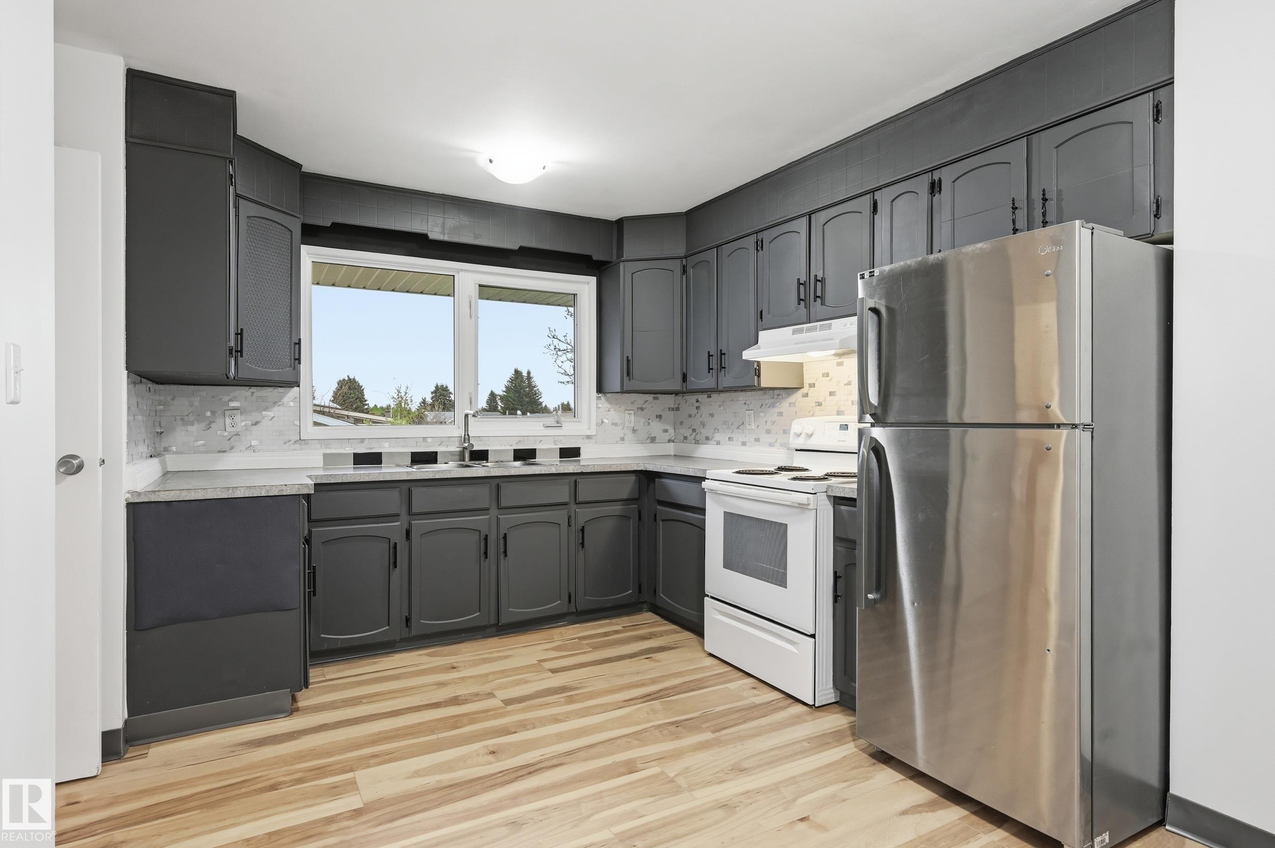Kitchen with dark cabinetry, light-toned wood-finish flooring, a stainless steel refrigerator, white electric range, and a dual-basin sink under a large window - 13903 87 Street, Edmonton, AB - Indoor Photo Showing Kitchen With Stainless Steel Kitchen With Double Sink