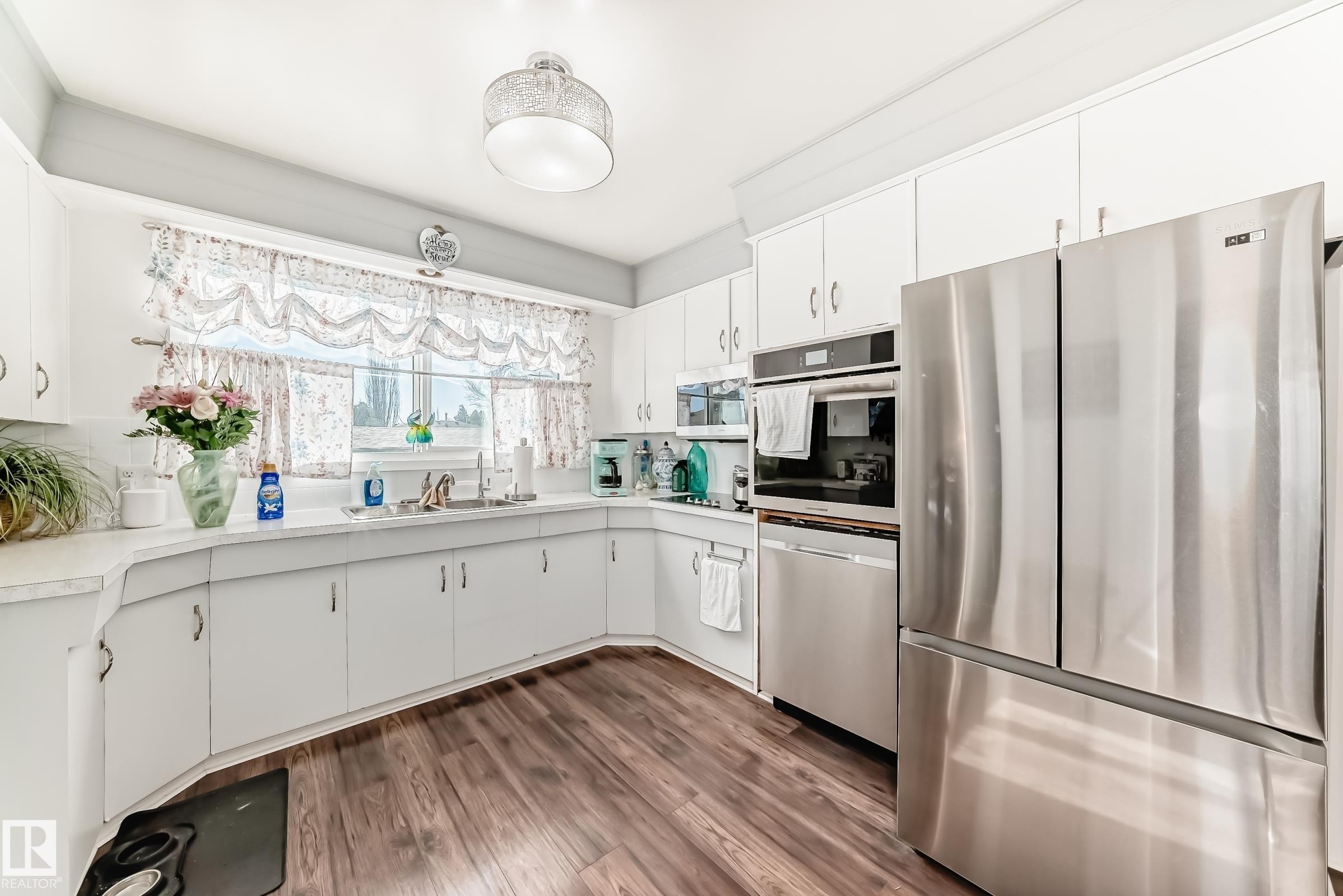 Bright kitchen featuring extensive white cabinetry, durable wood-finish flooring, a stainless steel French door refrigerator, built-in stainless steel oven, and a double basin sink beneath a large window - 3611 108 Street, Edmonton, AB - Indoor Photo Showing Kitchen With Stainless Steel Kitchen