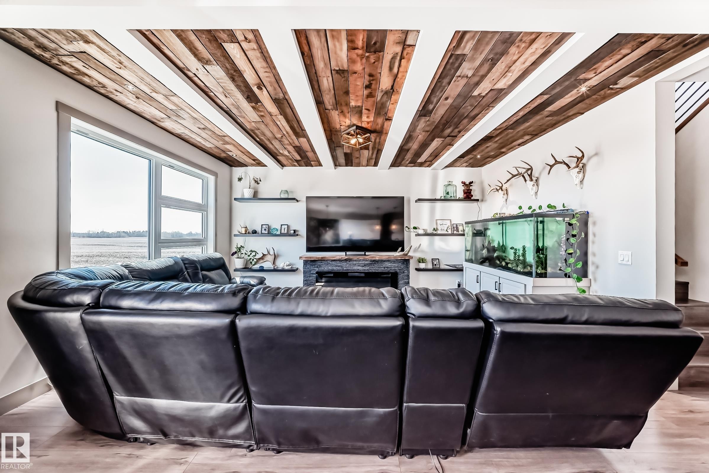 Living area featuring a distinctive wood-panel ceiling design, a built-in fireplace, wall-mounted shelving, and a large window - 5263 47 Ave, Calmar, AB - Indoor Photo Showing Living Room With Fireplace
