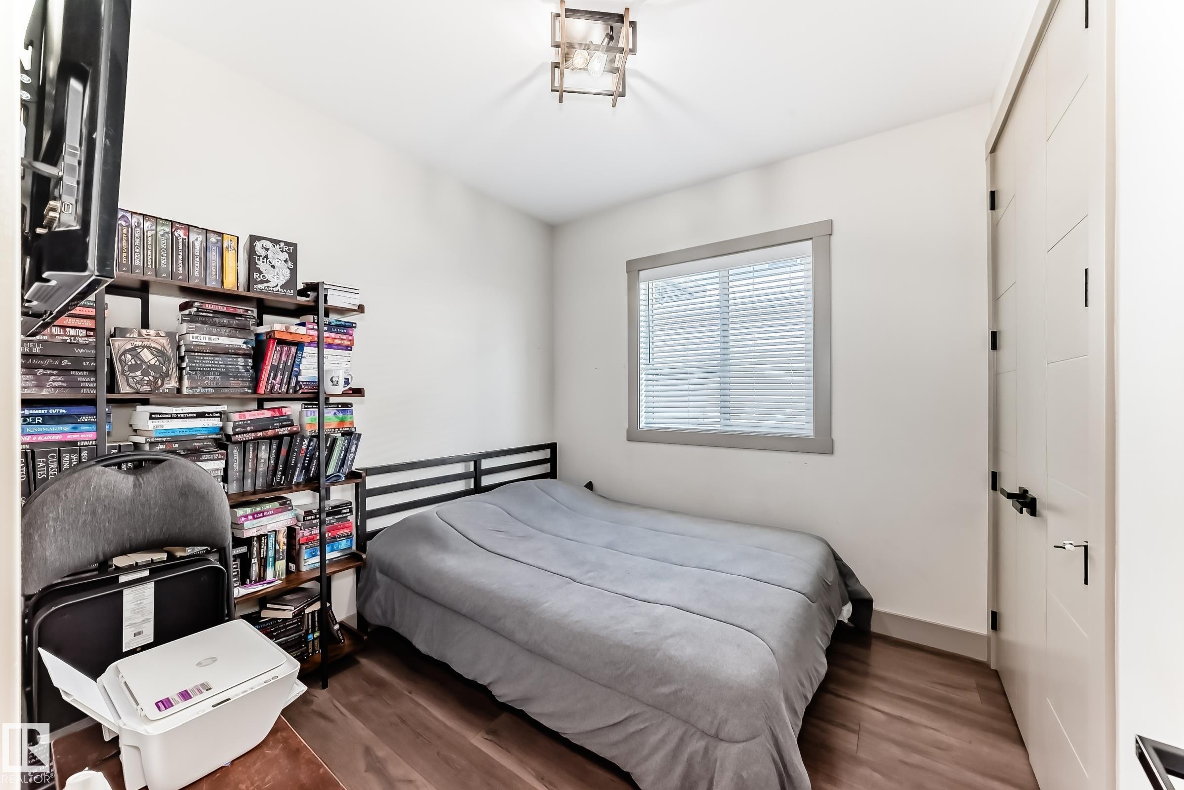 Bright interior space featuring wood-finish flooring, a contemporary ceiling light fixture, a window with horizontal blinds, and a modern interior door with black hardware - 5263 47 Ave, Calmar, AB - Indoor Photo Showing Bedroom