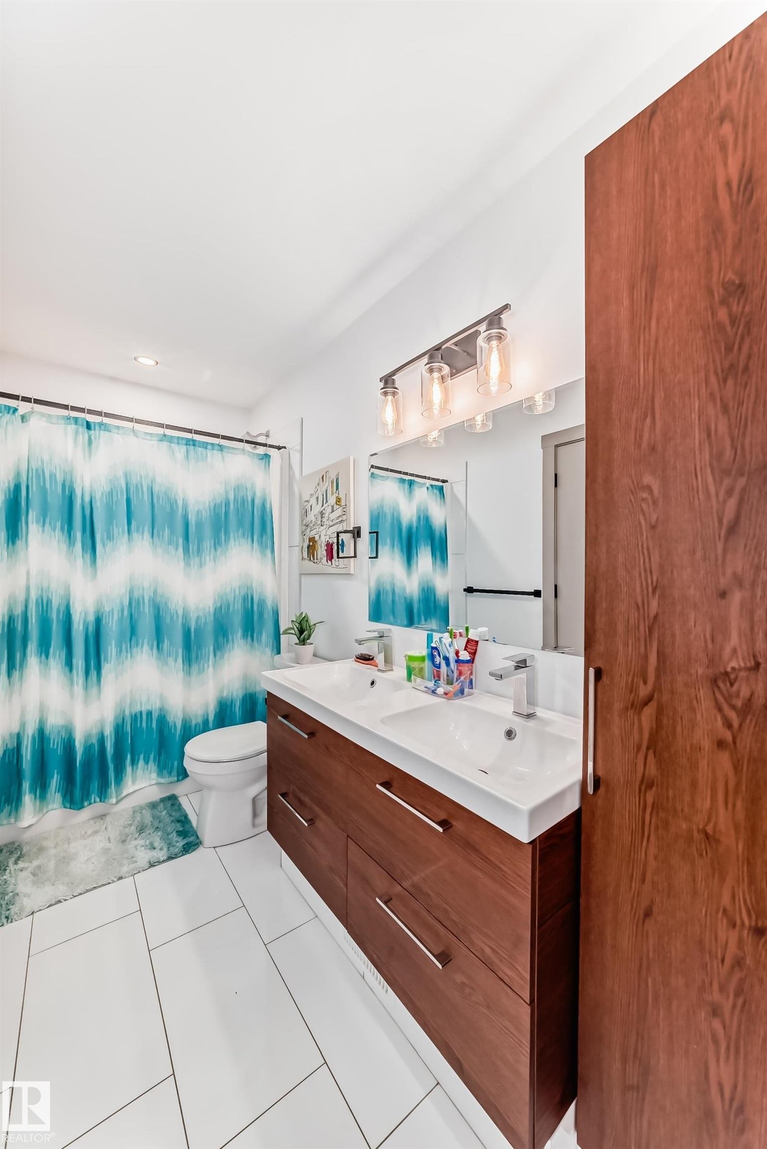Bathroom featuring a dual-sink vanity with wood-finish cabinetry, a large wall-mounted mirror, and white tile flooring - 5263 47 Ave, Calmar, AB - Indoor Photo Showing Bathroom