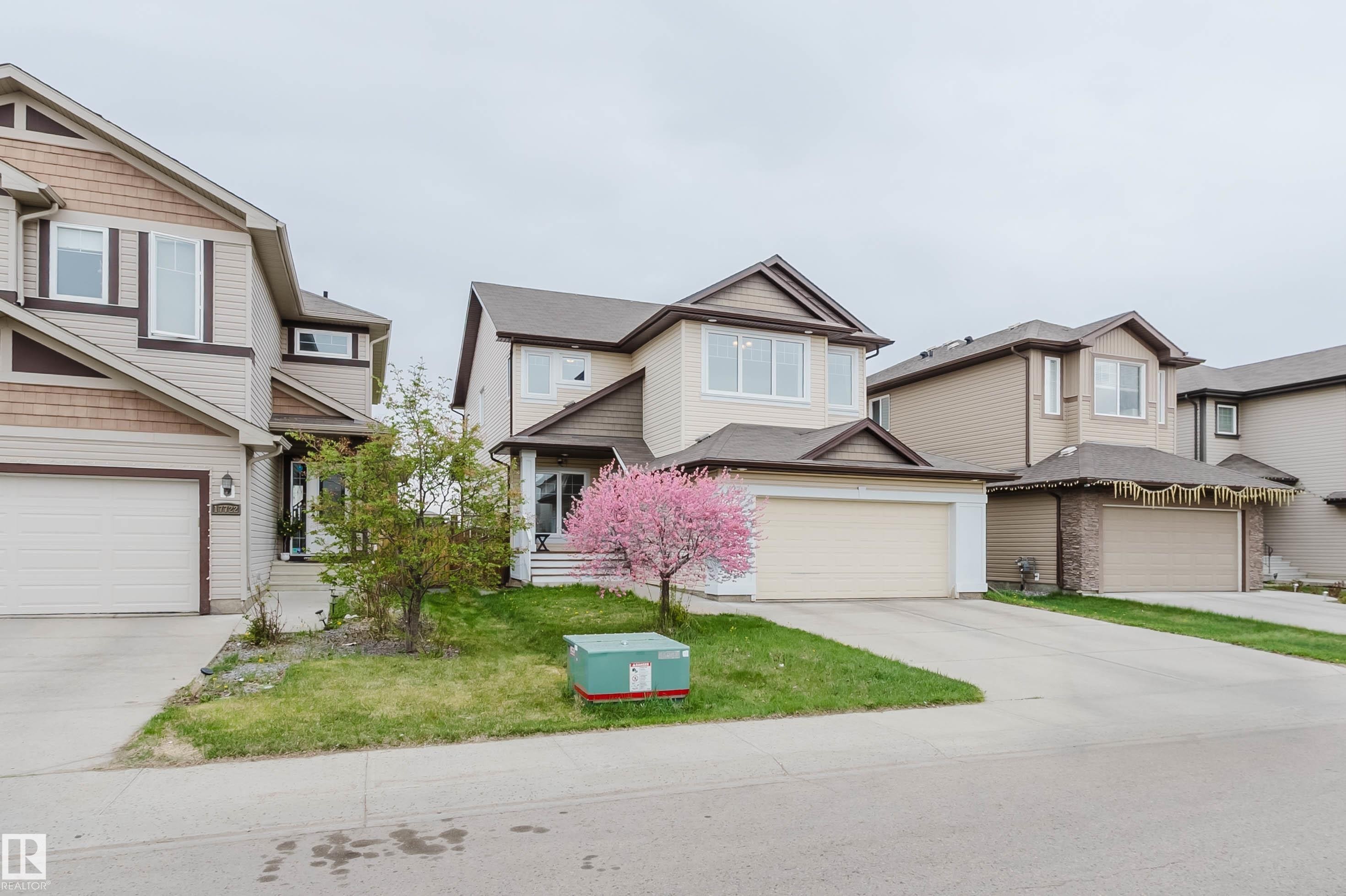 Two-story residence featuring light-toned siding with contrasting brown trim, an integrated garage, a paved driveway, and a front yard with a blooming tree - 17718 5A Avenue, Edmonton, AB - Outdoor With Facade