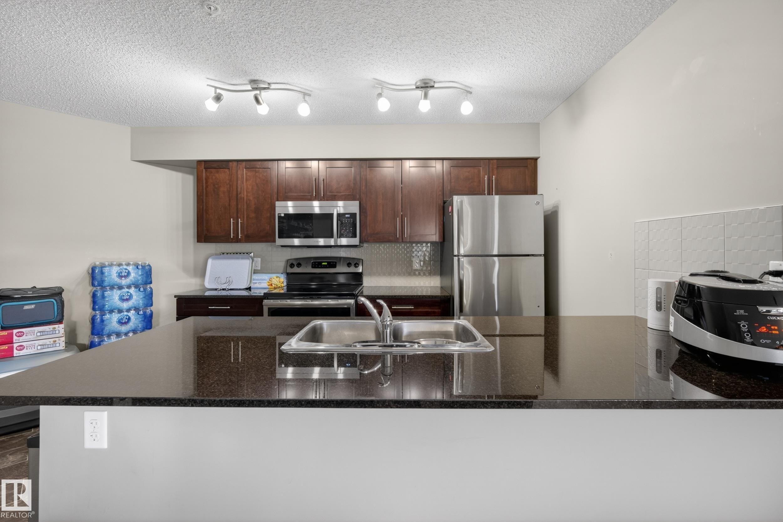 Kitchen featuring dark wood-finish cabinetry, stainless steel appliances, a dark countertop, and track lighting fixtures - 314 3211 James Mowatt Trail Sw, Edmonton, AB - Indoor Photo Showing Kitchen With Stainless Steel Kitchen With Double Sink