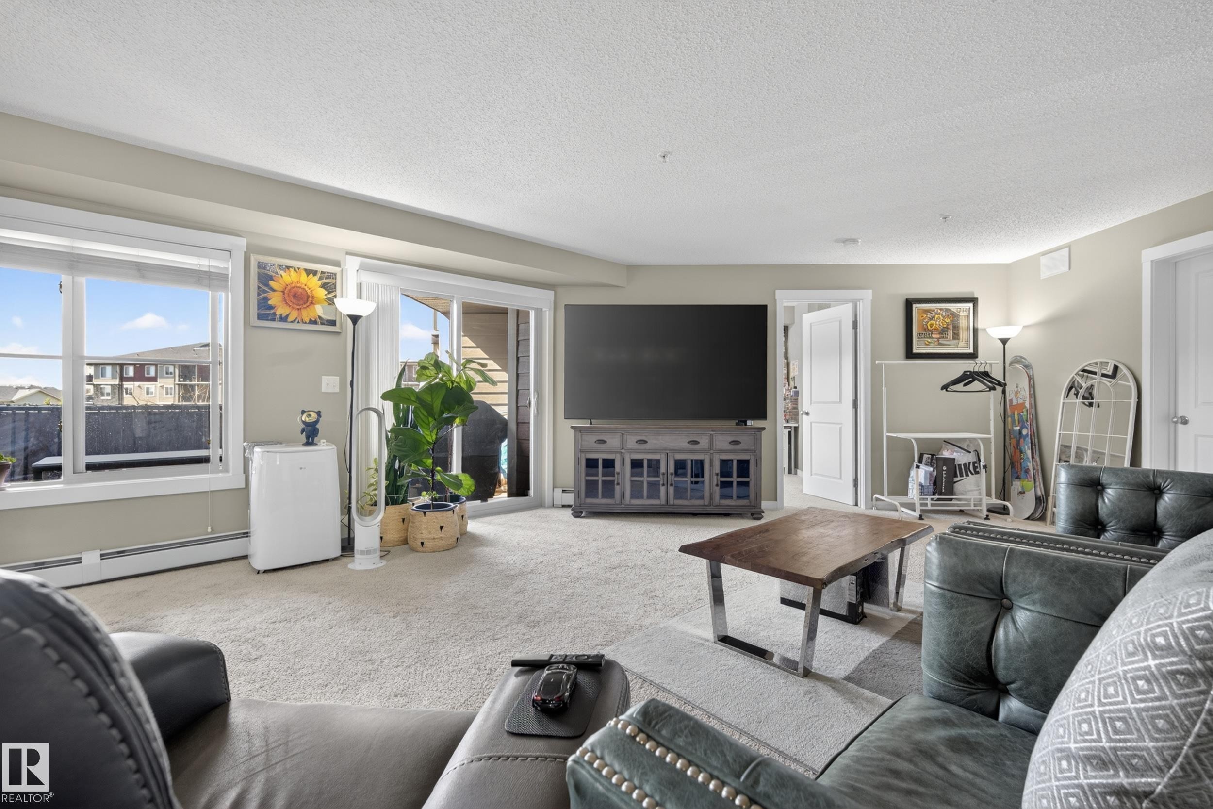Carpeted living area featuring a large window, sliding glass door, white baseboards, and neutral wall tones - 314 3211 James Mowatt Trail Sw, Edmonton, AB - Indoor Photo Showing Living Room