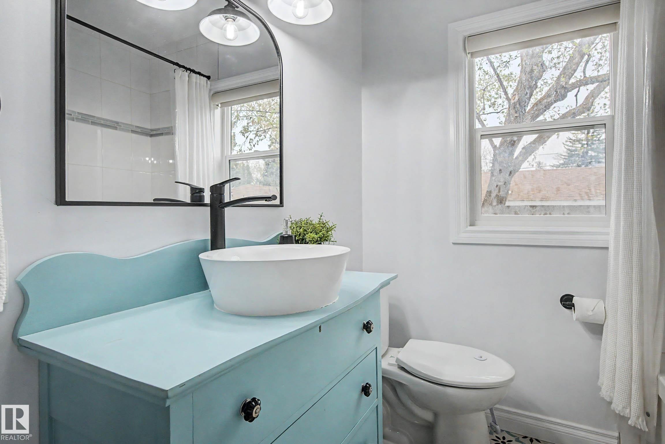 Bathroom featuring a light blue vanity with a vessel sink and matte black faucet, a large rectangular mirror with a metal frame, and a window with white trim - 13316 109A Avenue, Edmonton, AB - Indoor Photo Showing Bathroom