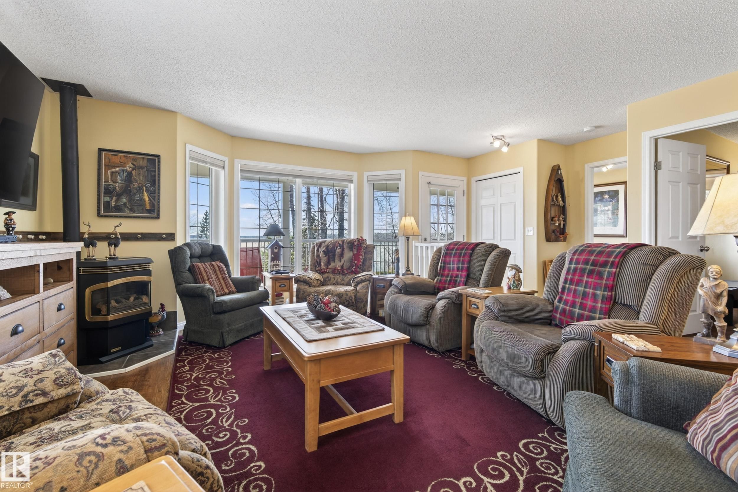 Living area featuring a bay window with multiple panes, a built-in wood-burning stove, light-colored walls, and a patterned area rug - 77 Lakeshore Drive, Rural Leduc County, AB - Indoor Photo Showing Living Room With Fireplace