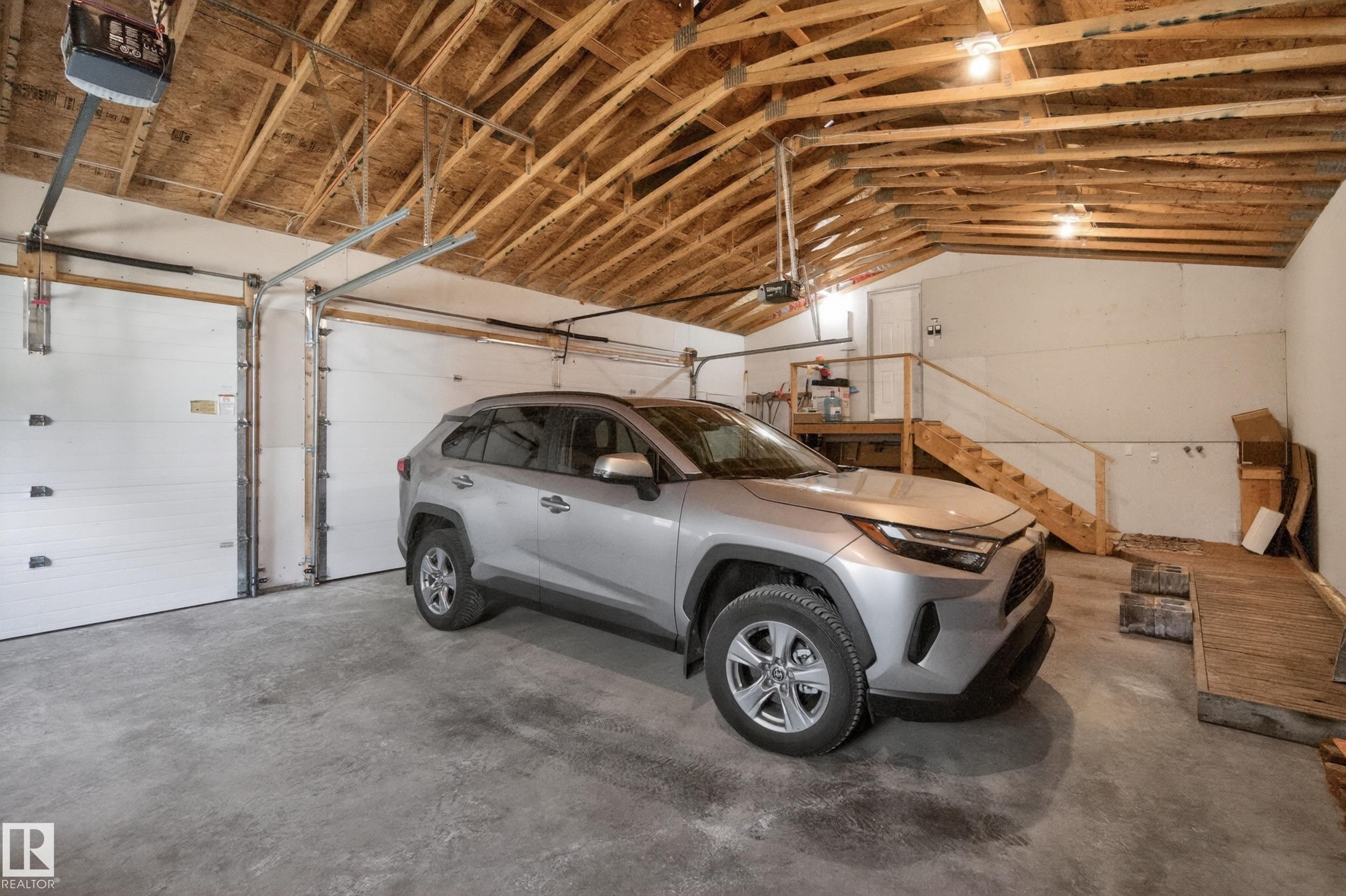 Two-car garage featuring exposed wood rafters, concrete flooring, dual automatic garage door openers, and a built-in wooden staircase - 77 Lakeshore Drive, Rural Leduc County, AB - Indoor Photo Showing Garage
