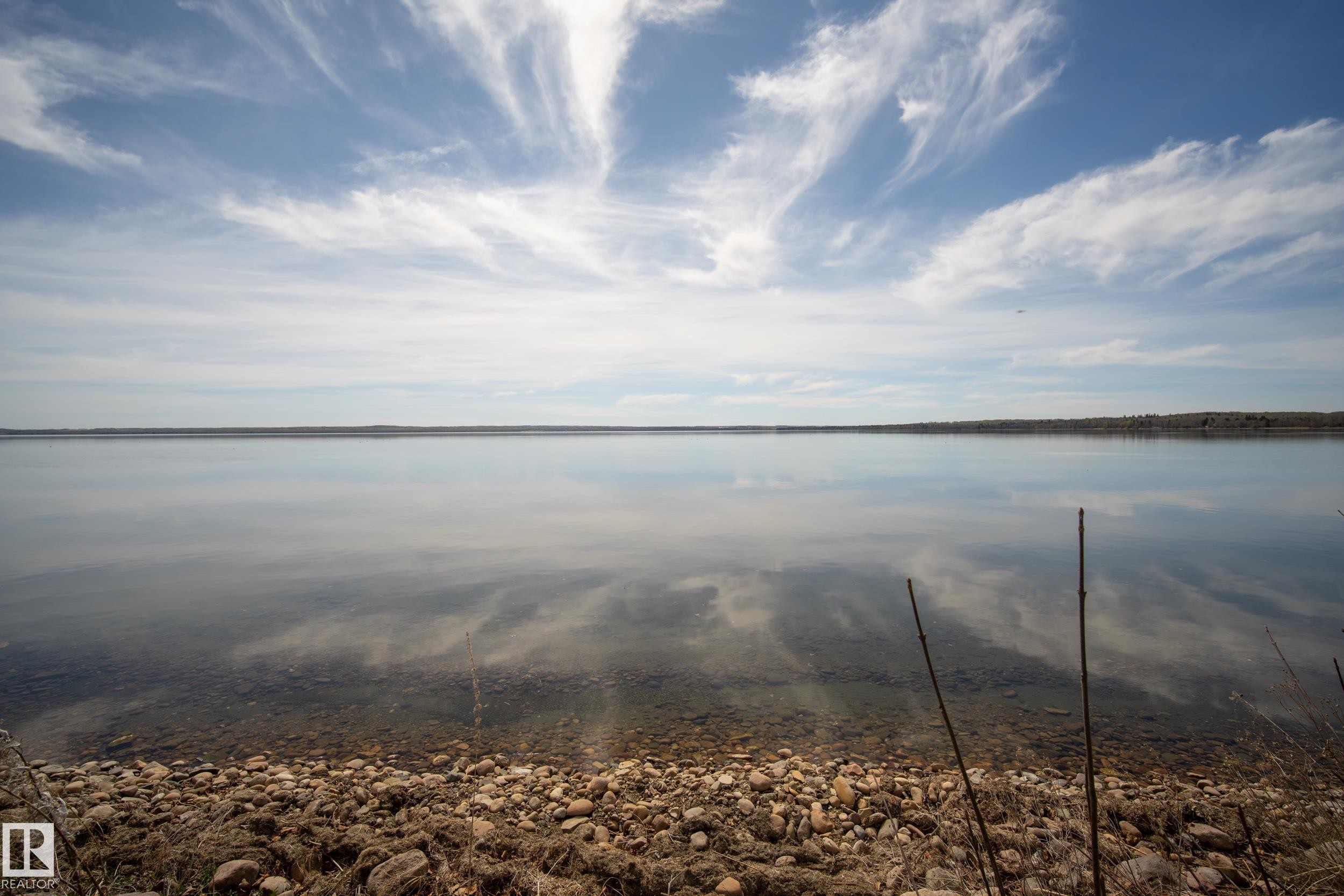 Tranquil waterscape featuring a rocky shoreline and a reflective surface mirroring the sky - 77 Lakeshore Drive, Rural Leduc County, AB - Outdoor With Body Of Water With View