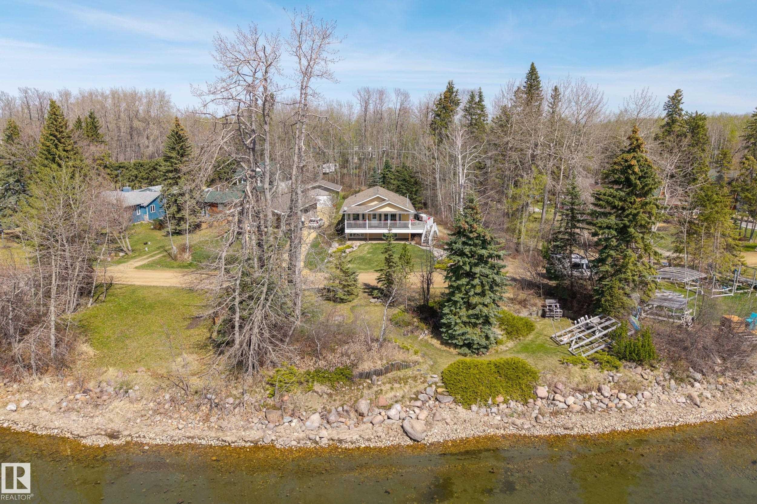 Waterfront property featuring a yellow exterior with white trim, a spacious deck with white railings, mature evergreen trees, a gently sloping lawn, and a natural shoreline - 77 Lakeshore Drive, Rural Leduc County, AB - Outdoor With Body Of Water With View
