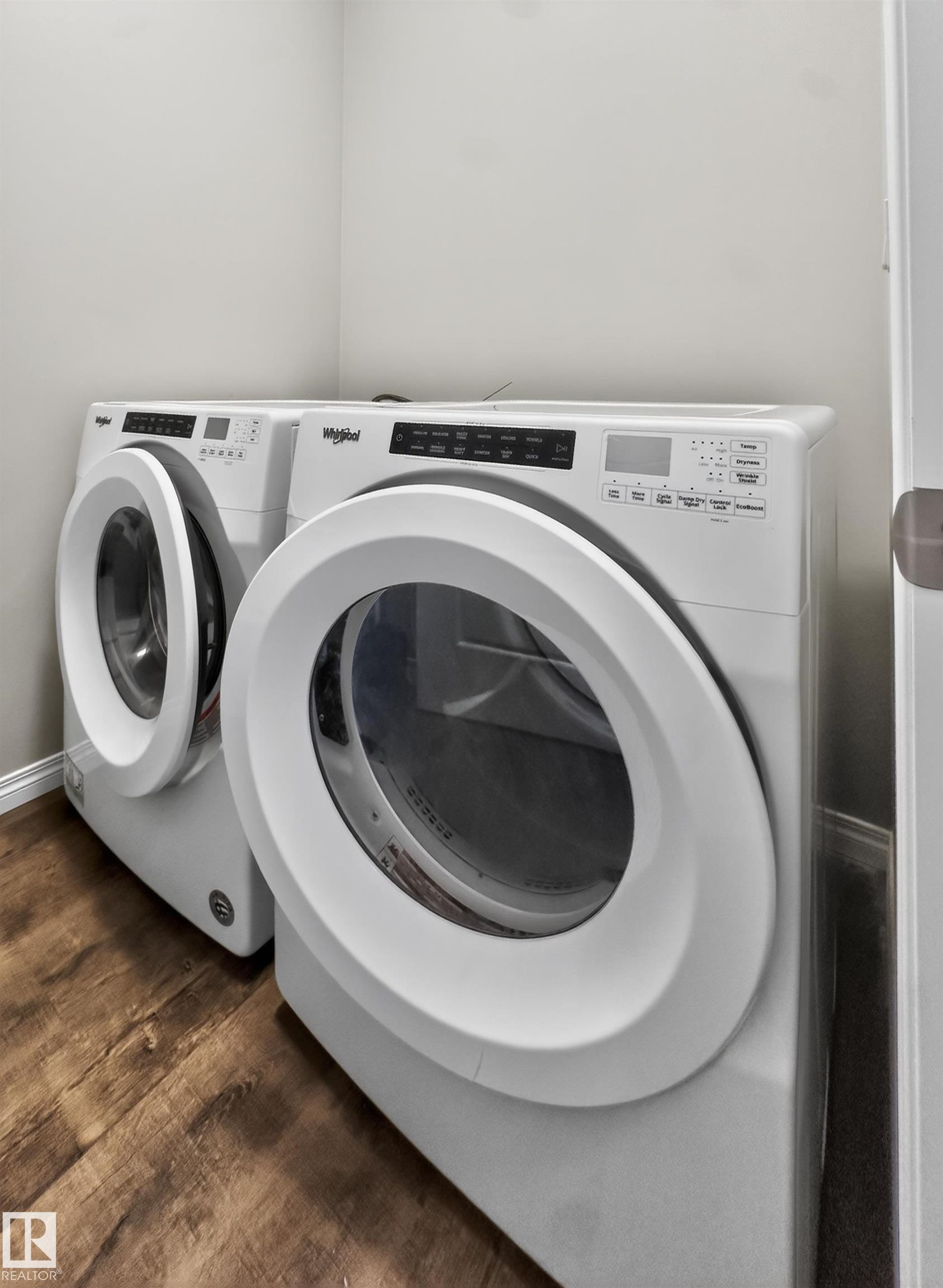 A dedicated laundry area featuring a front-loading washing machine and dryer, set against light-colored walls and wood-look flooring - 15 2072 Wonnacott Way, Edmonton, AB - Indoor Photo Showing Laundry Room