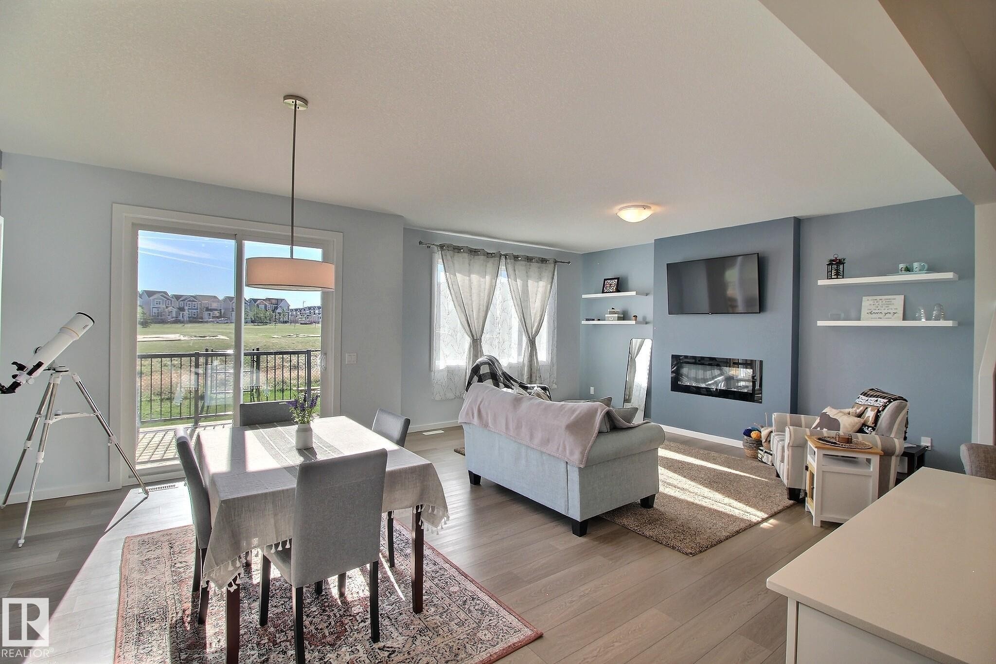 Open-concept living area featuring light wood-finish flooring, a built-in electric fireplace, and a sliding glass door leading to an outdoor balcony with metal railings - 121 Stiles Bend, Leduc, AB - Indoor With Fireplace