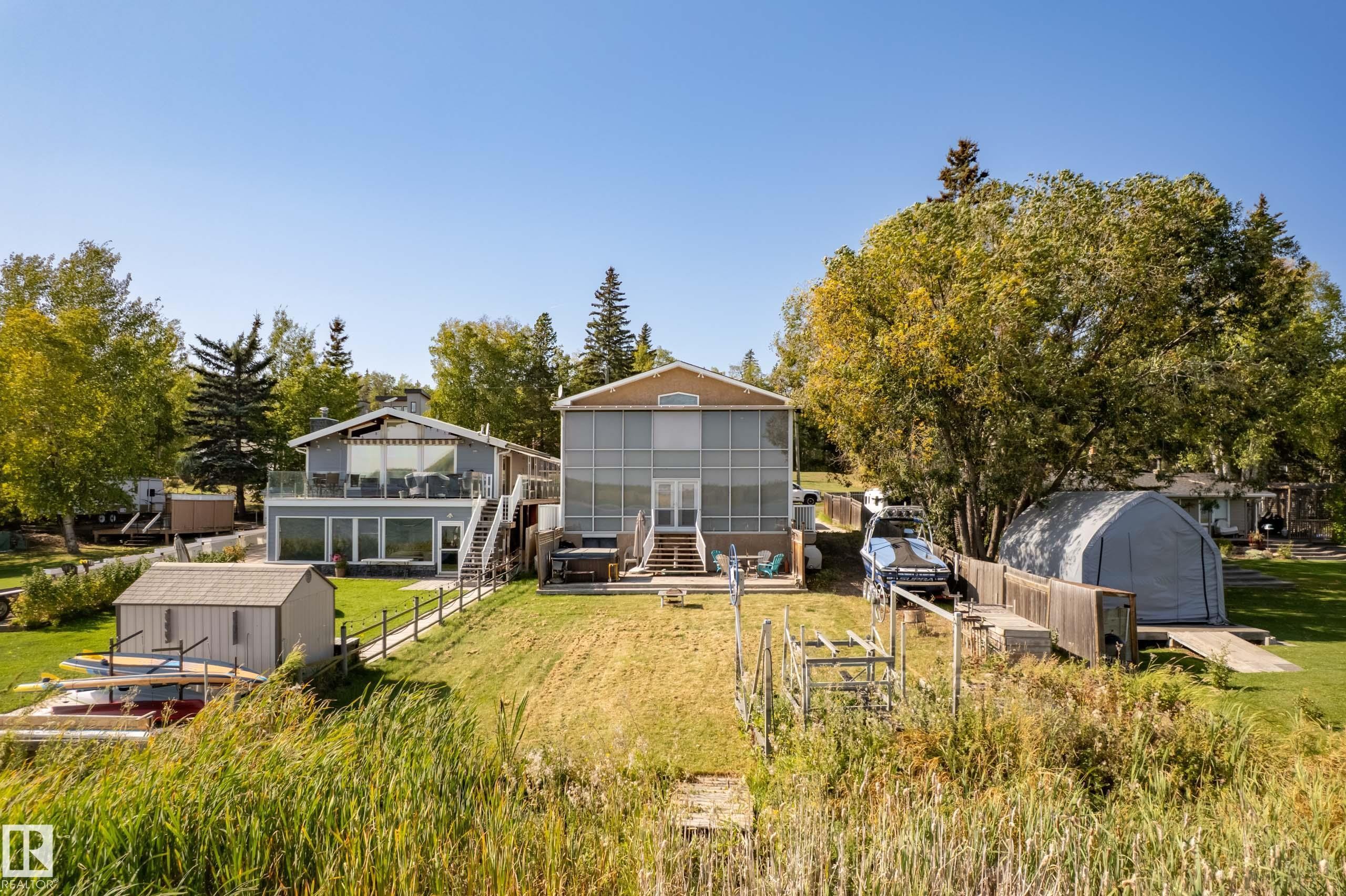 Waterfront property featuring two structures, established landscaping, a ground-level patio, an upper-level deck with glass railings, and a detached storage shed - 104 Bayview Drive, Rural Parkland County, AB - Outdoor With Deck Patio Veranda
