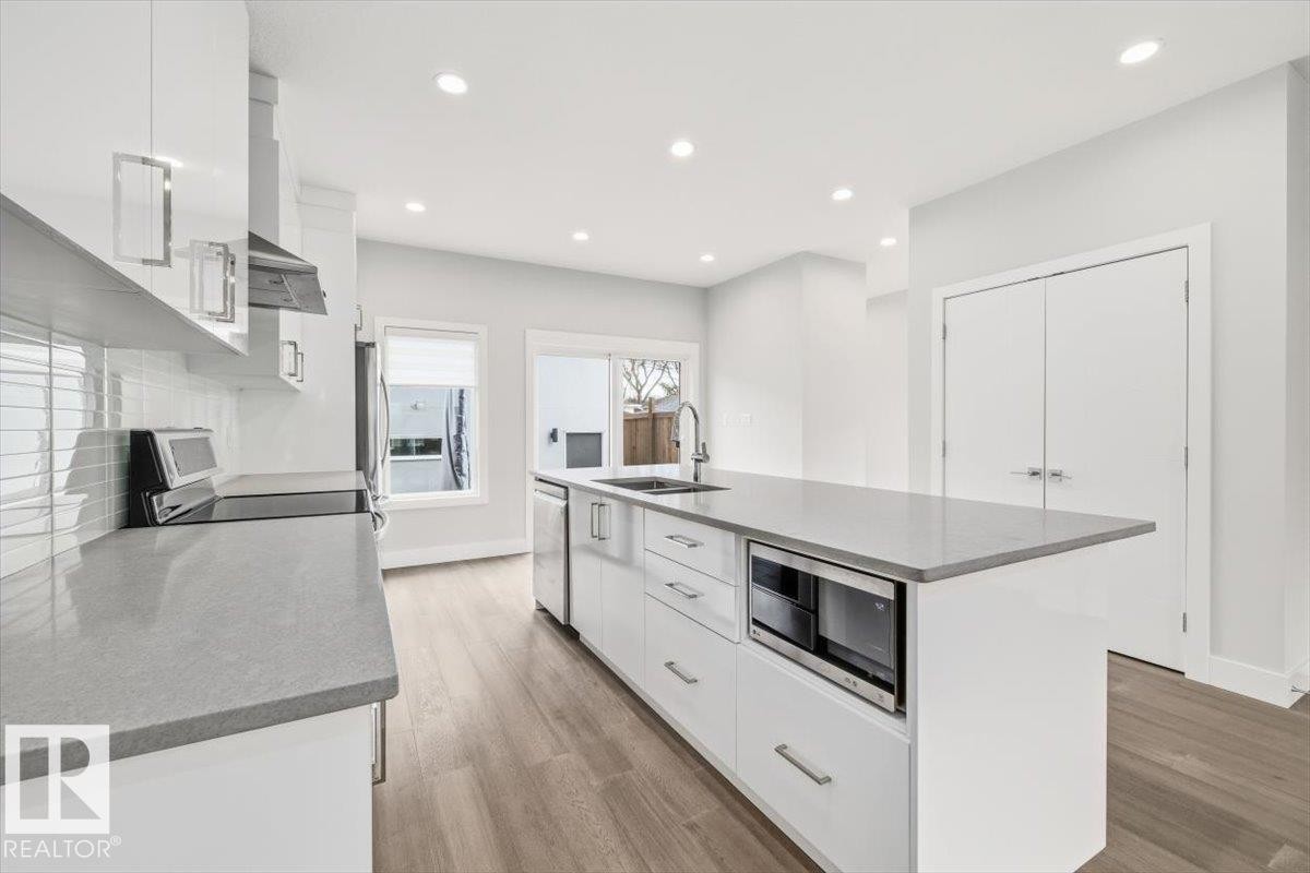 Open-concept kitchen featuring an expansive center island with an under-counter microwave, crisp white cabinetry, light-toned wood-finish flooring, recessed lighting, and stainless steel appliances - 9933 79 Street, Edmonton, AB - Indoor Photo Showing Kitchen With Double Sink