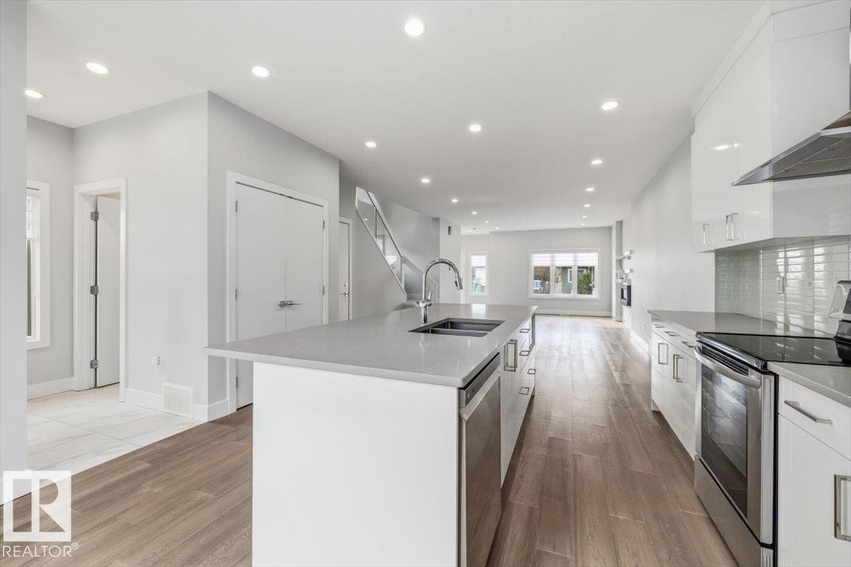Modern kitchen featuring a large island with an undermount sink, stainless steel appliances, white flat-panel cabinetry, and wood-finish flooring - 9933 79 Street, Edmonton, AB - Indoor Photo Showing Kitchen With Double Sink With Upgraded Kitchen