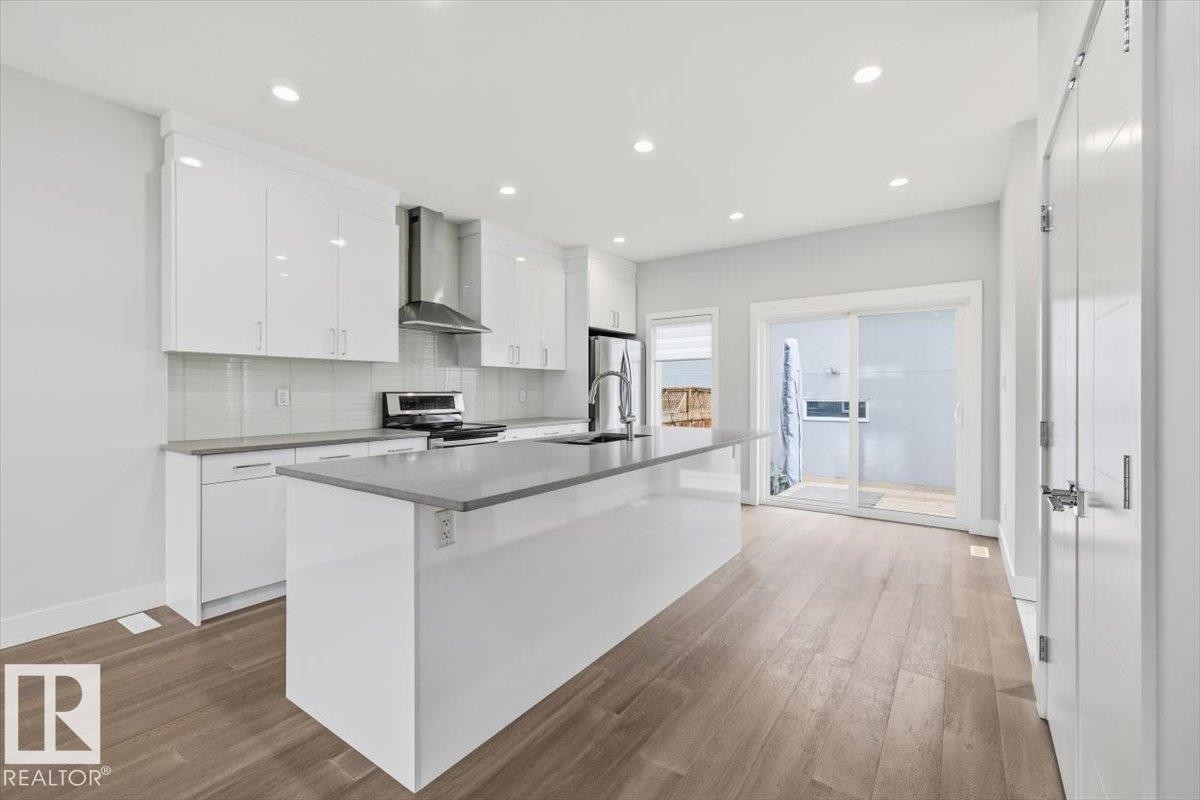 Contemporary kitchen featuring a large island, white cabinetry, integrated stainless steel appliances, wood-finish flooring, and sliding glass door access to an outdoor area - 9933 79 Street, Edmonton, AB - Indoor Photo Showing Kitchen With Upgraded Kitchen