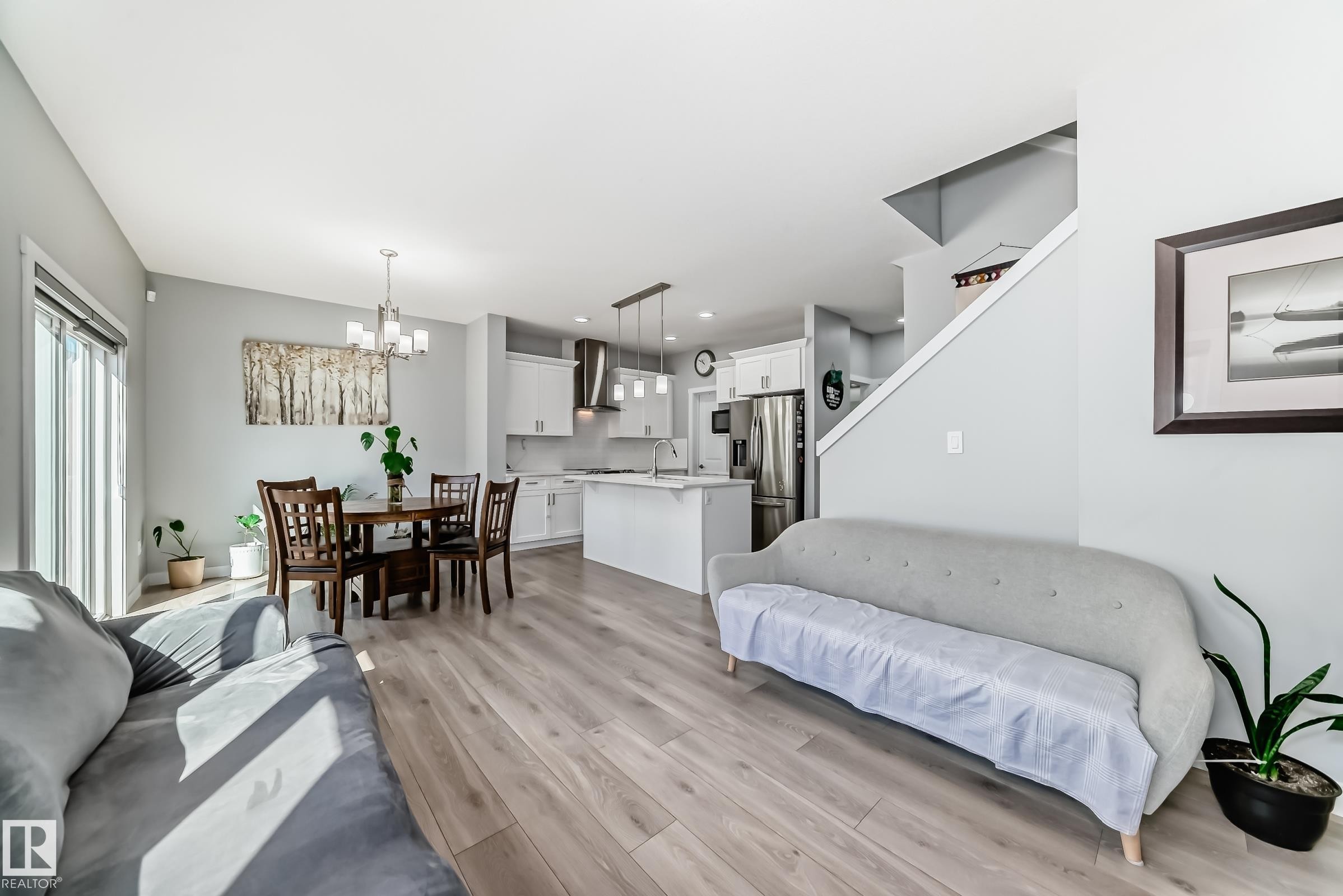 Open-concept interior featuring wood-finish flooring, a kitchen with white cabinetry and stainless steel appliances, a dining area with a contemporary chandelier, and sliding glass doors - 19635 29 Avenue, Edmonton, AB - Indoor