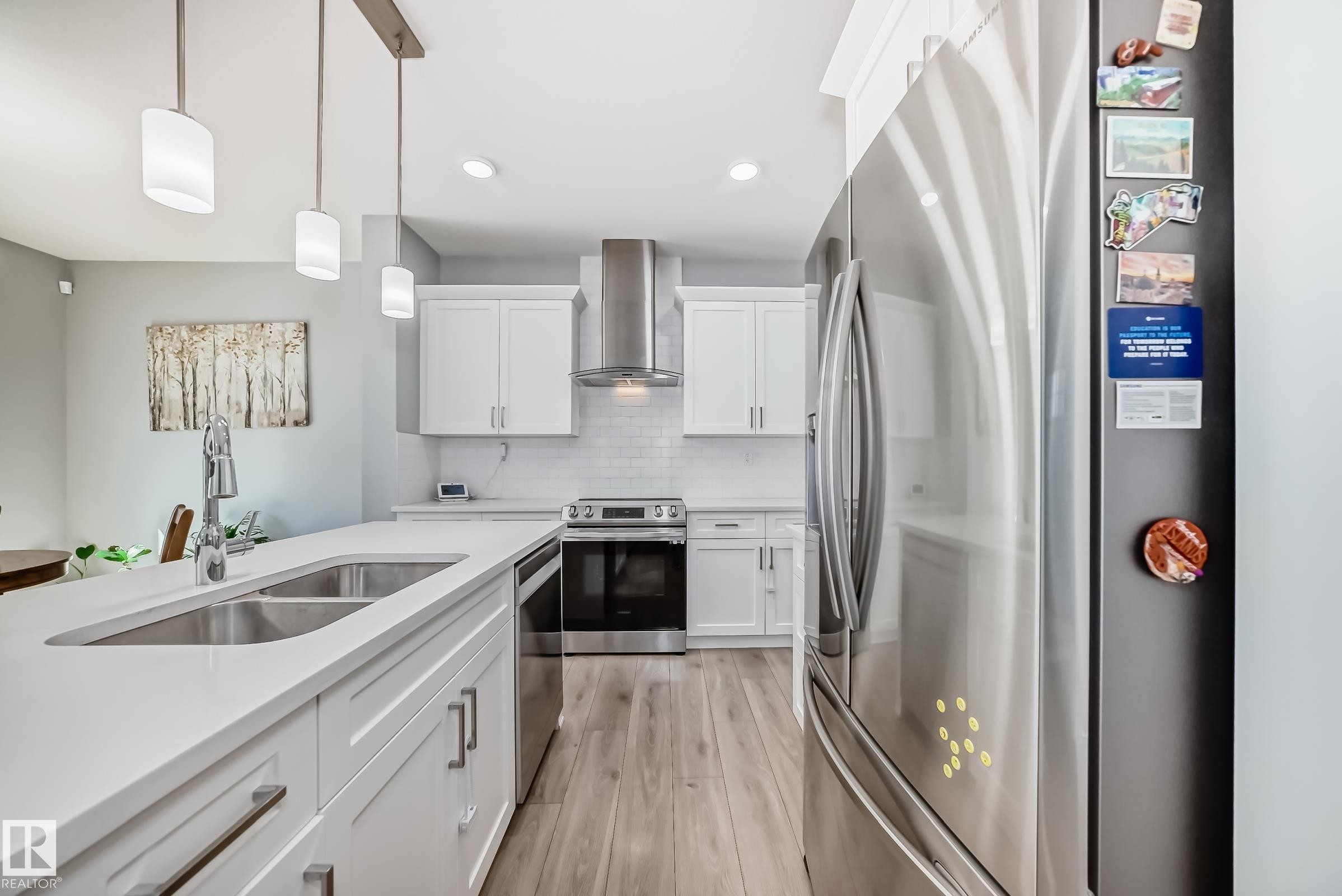 Modern kitchen featuring a large island with an undermount double sink, white cabinetry, integrated stainless steel appliances, wood-finish flooring, and pendant lighting - 19635 29 Avenue, Edmonton, AB - Indoor Photo Showing Kitchen With Double Sink With Upgraded Kitchen