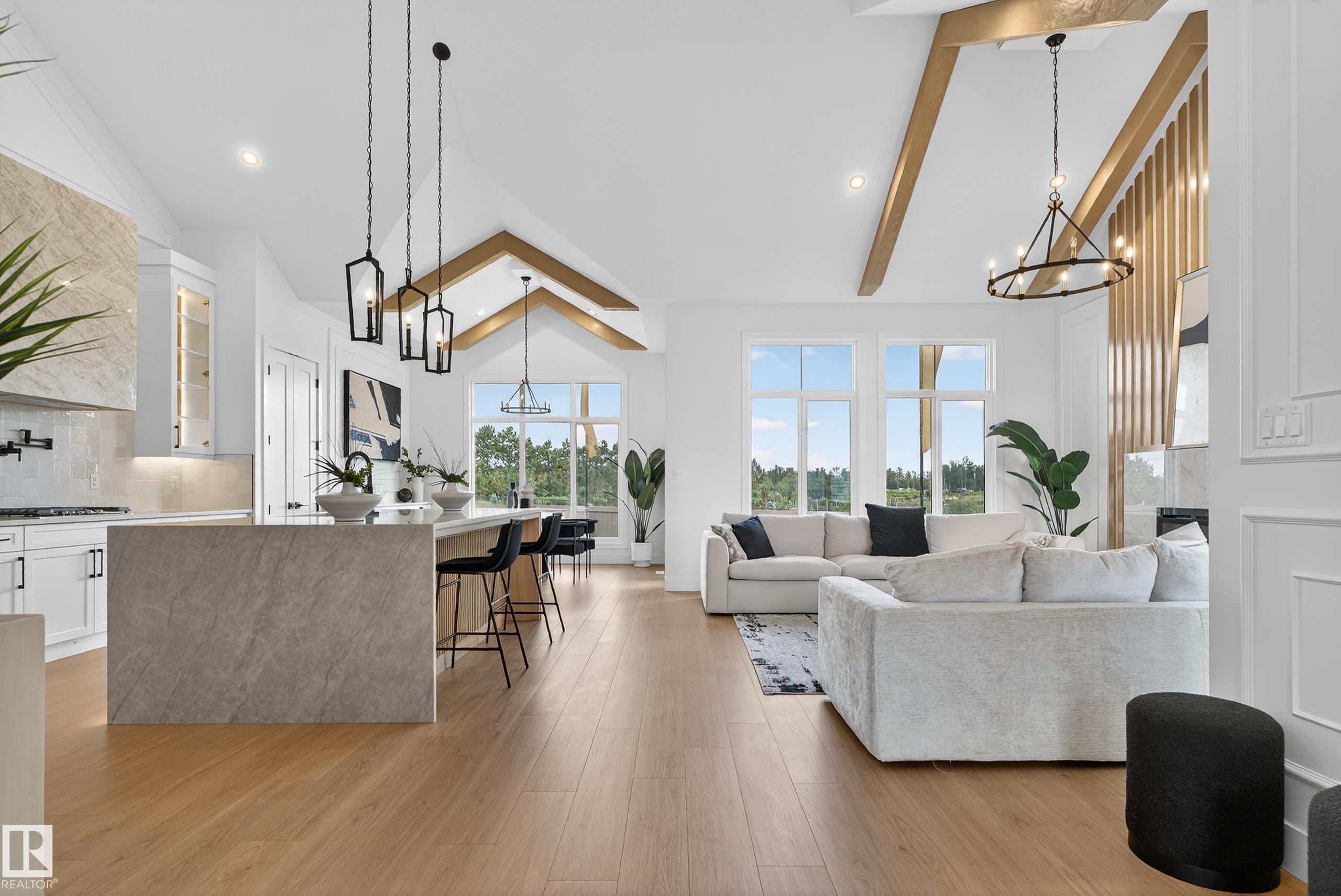 Open-concept living area featuring a vaulted ceiling with exposed beams, wood-finish flooring, a kitchen island with seating, a tile backsplash, and large windows - 711 166 Avenue, Edmonton, AB - Indoor