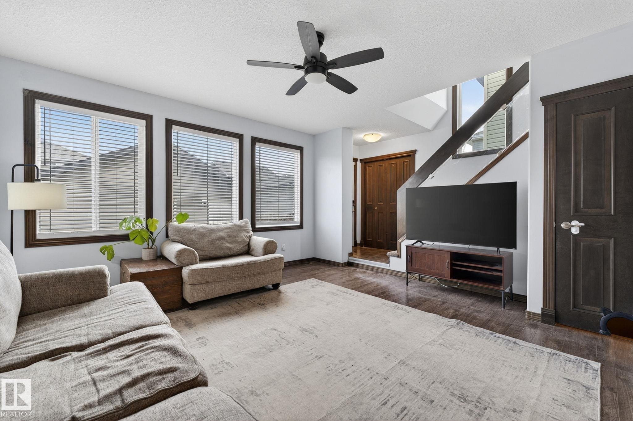 Living area featuring dark wood-finish flooring, three large windows with blinds, a dark ceiling fan, a built-in staircase, and a dark wood door - 108 Hewitt Circle, Spruce Grove, AB - Indoor Photo Showing Living Room