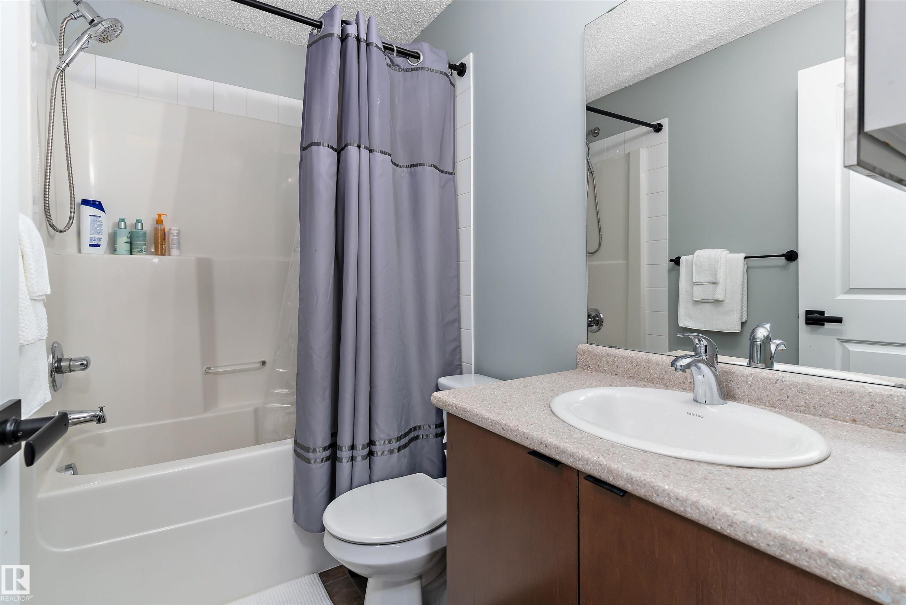 Bathroom featuring a combination shower and tub with white surround, a toilet, and a vanity with an integrated sink and wood-finish cabinetry - 122 3357 16A Avenue, Edmonton, AB - Indoor Photo Showing Bathroom