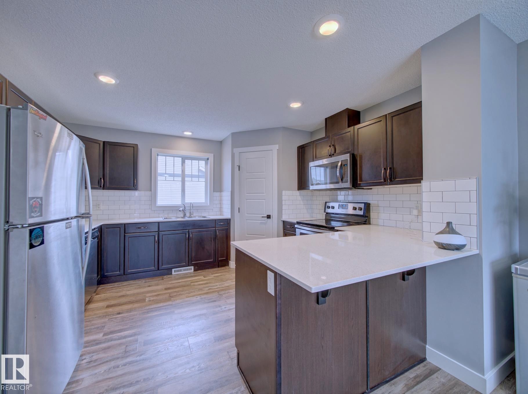 Modern kitchen featuring a central island with a white countertop, dark wood-finish cabinetry, stainless steel appliances, white subway tile backsplash, and recessed lighting - 11 165 Cy Becker Boulevard, Edmonton, AB - Indoor Photo Showing Kitchen
