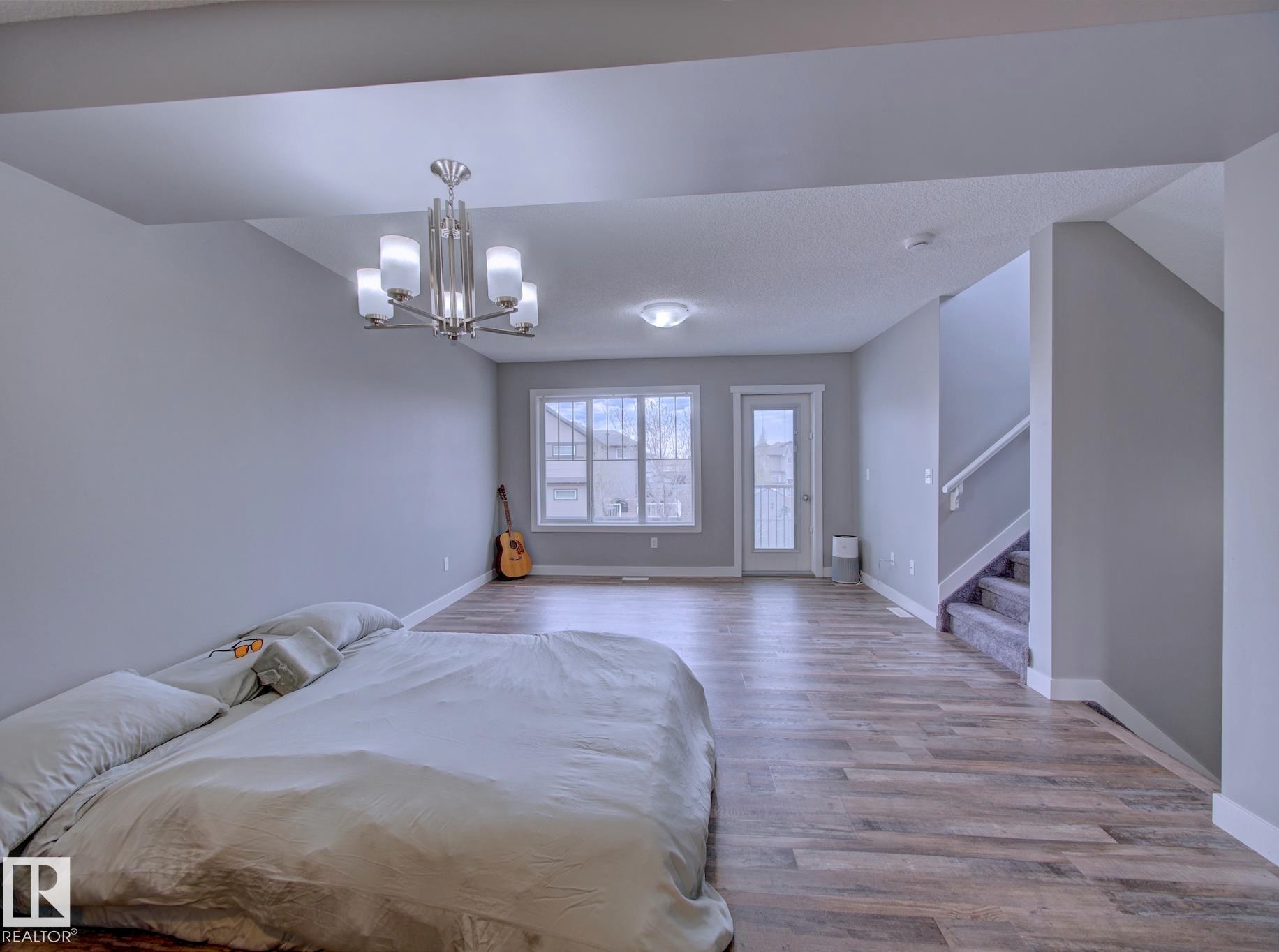 Spacious room featuring wood-finish flooring, a contemporary chandelier, large windows, a glass-paneled door, and a carpeted staircase with white railings - 11 165 Cy Becker Boulevard, Edmonton, AB - Indoor Photo Showing Bedroom