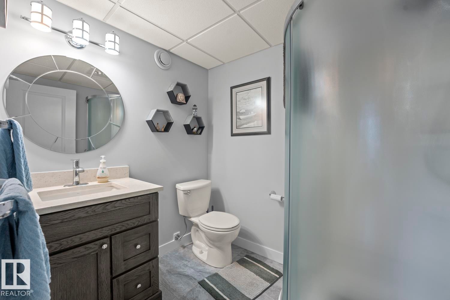 Bathroom featuring a dark wood-finish vanity with a light-toned countertop and an undermount sink, a large decorative circular mirror, a three-light vanity fixture, a toilet, and a frosted glass shower enclosure - 1304 12 Street, Cold Lake, AB - Indoor Photo Showing Bathroom