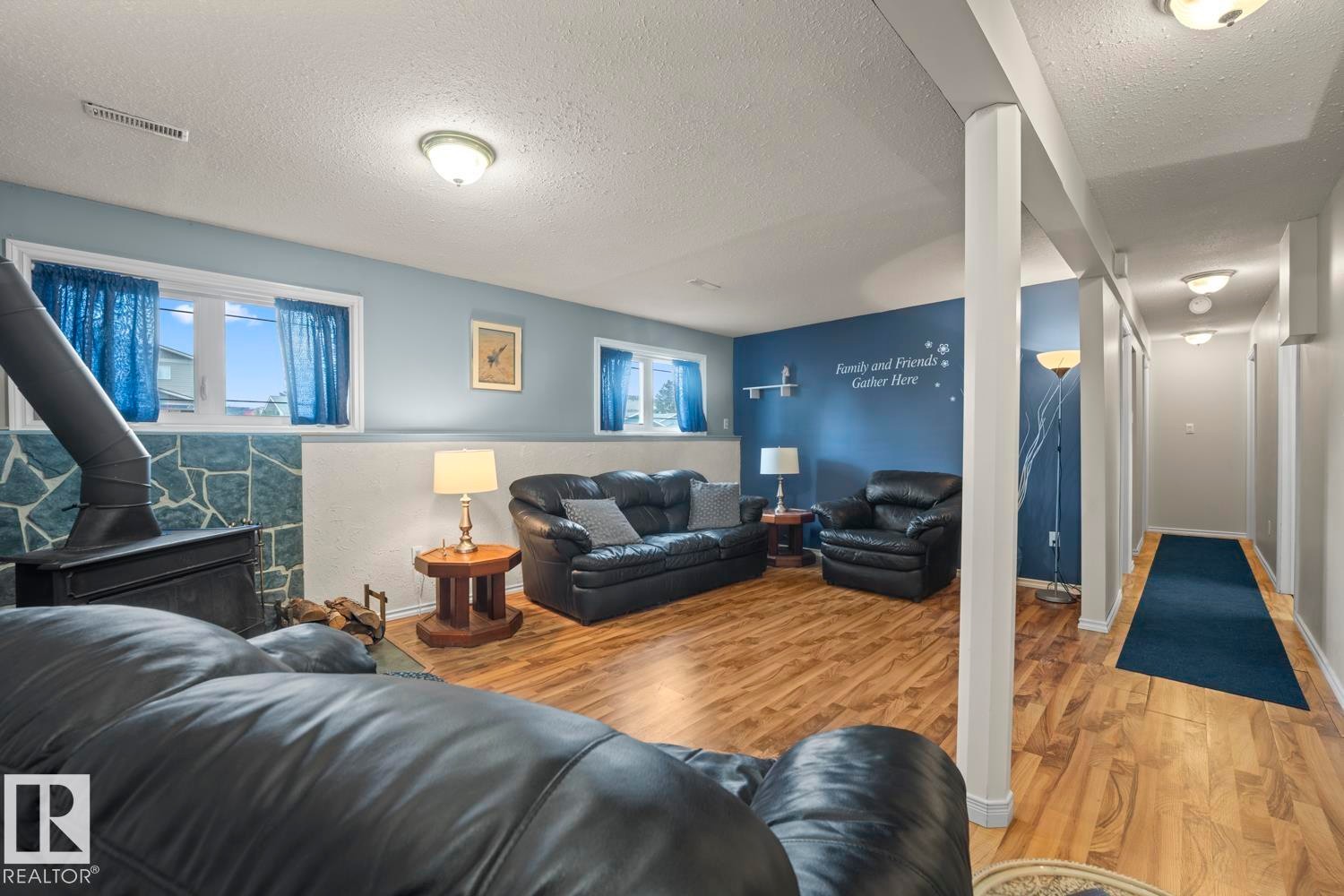 Spacious recreation room featuring wood-finish flooring, a stone-clad wall with a black wood-burning stove, and multiple windows - 1304 12 Street, Cold Lake, AB - Indoor Photo Showing Living Room