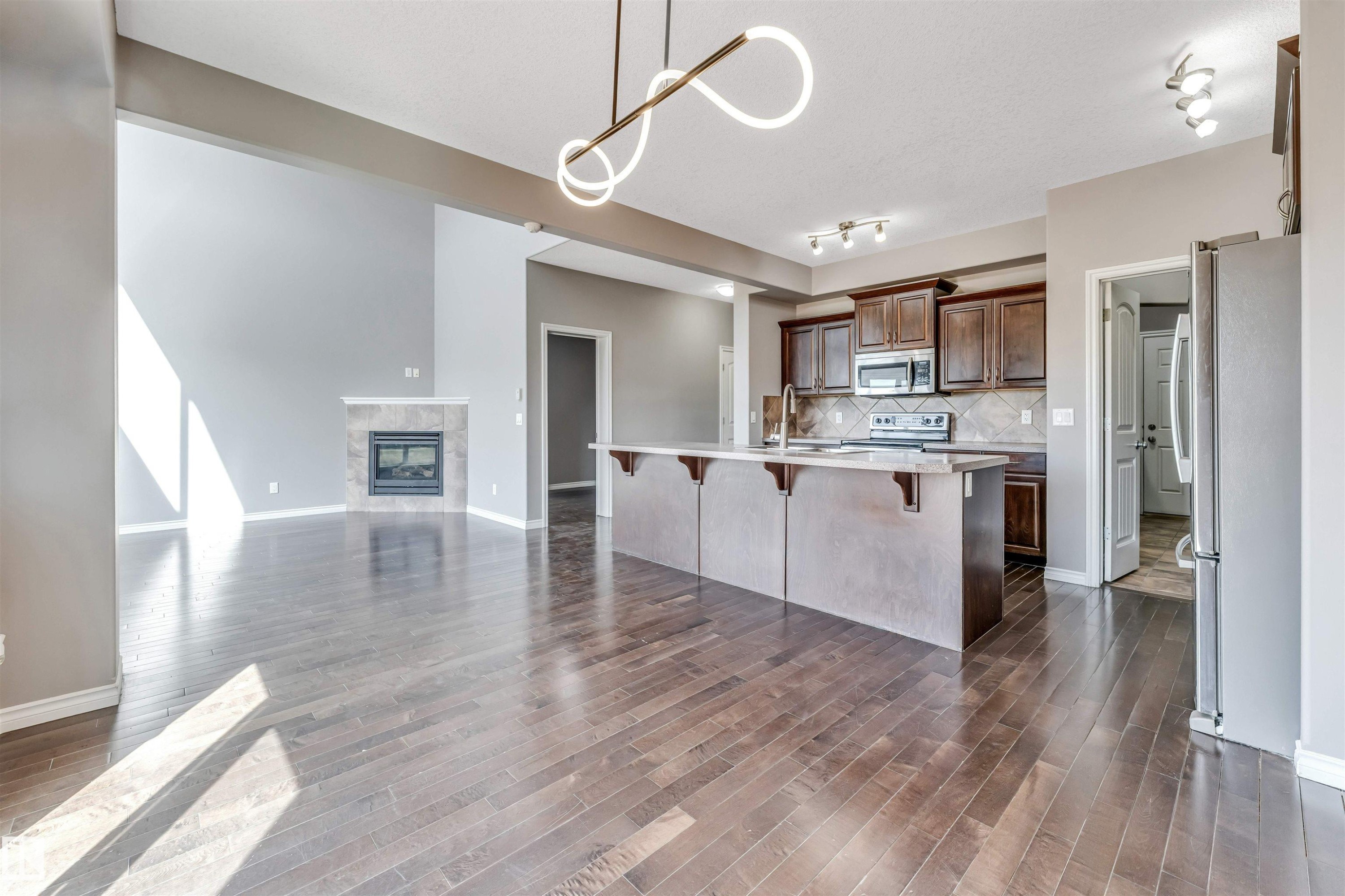 Open-concept living area featuring rich wood-finish flooring, a contemporary fireplace with a tiled surround, a large kitchen island with a breakfast bar, dark wood cabinetry, and stainless steel appliances - 2138 32B Street, Edmonton, AB - Indoor Photo Showing Kitchen