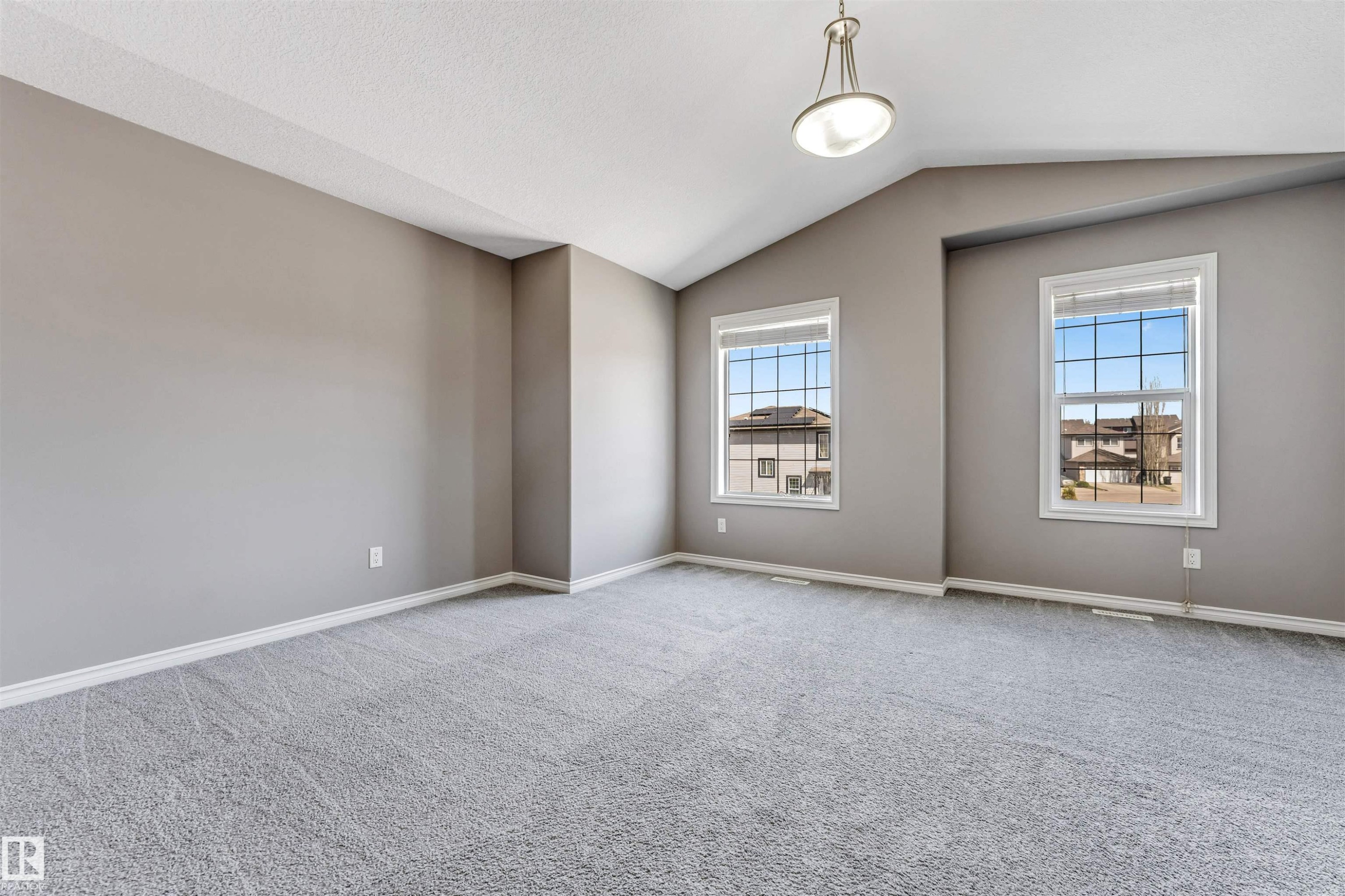 Spacious room featuring vaulted ceilings, a suspended light fixture, two double-paned windows with horizontal blinds, neutral wall paint, and gray patterned carpeting - 2138 32B Street, Edmonton, AB - Indoor Photo Showing Other Room